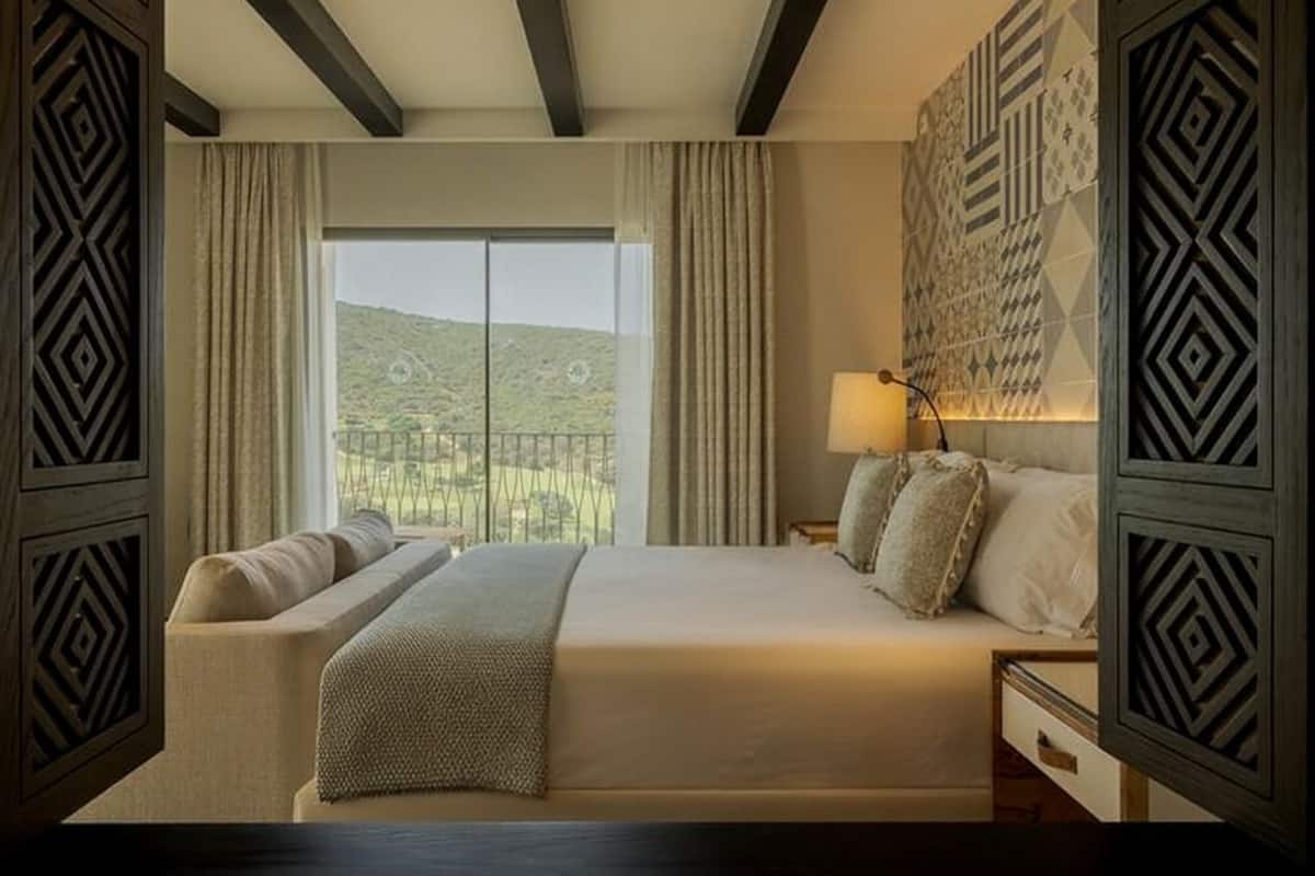 Une chambre avec un grand lit, une table de chevet, une lampe et une vue sur un paysage verdoyant par la fenêtre.