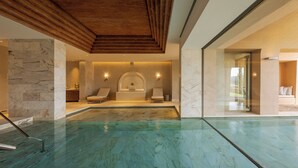 A modern indoor swimming pool with a wooden ceiling, marble walls, and a seating area with a bench and a small table.