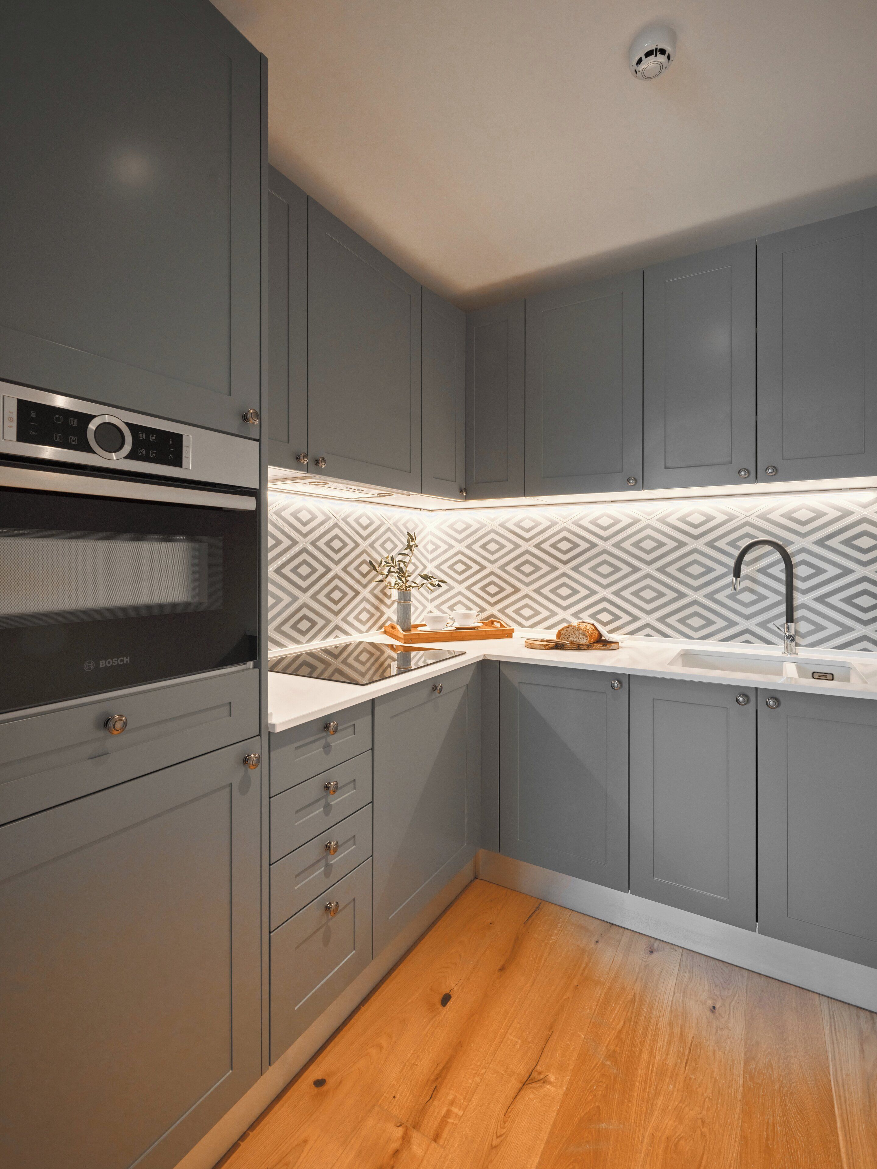 A modern kitchen with grey cabinets, a Bosch oven, a patterned backsplash, and a wooden floor.