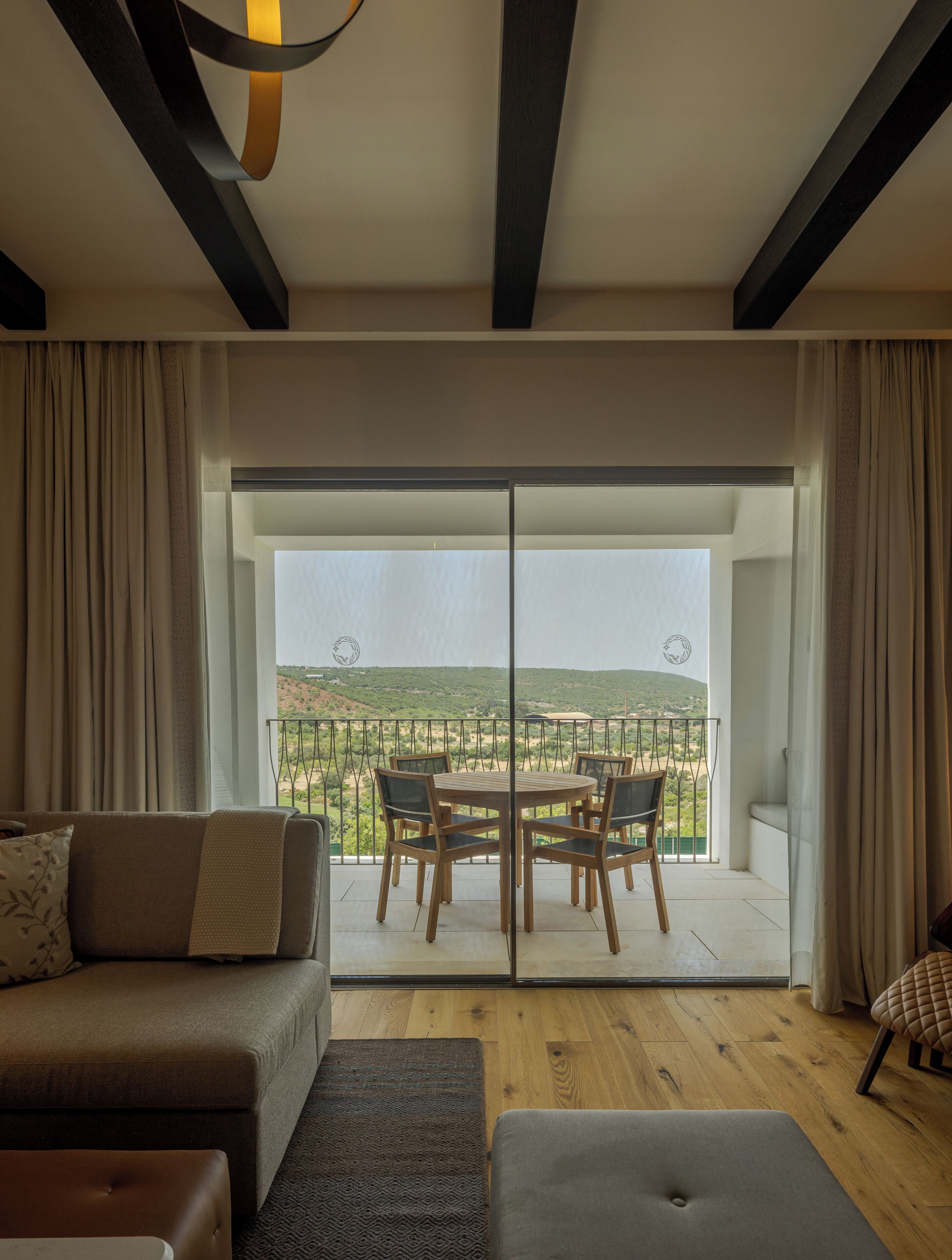 A modern living room with a sofa, a coffee table, and a dining area with chairs and a table. There is a large sliding glass door leading to a balcony with a view of the countryside.