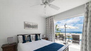 A bedroom with a bed, a nightstand, a ceiling fan, a window with curtains, and a view of the ocean. - 25 Westmoreland Hills | Modern Caribbean Villa with Spectacular Sunset Views (St James)