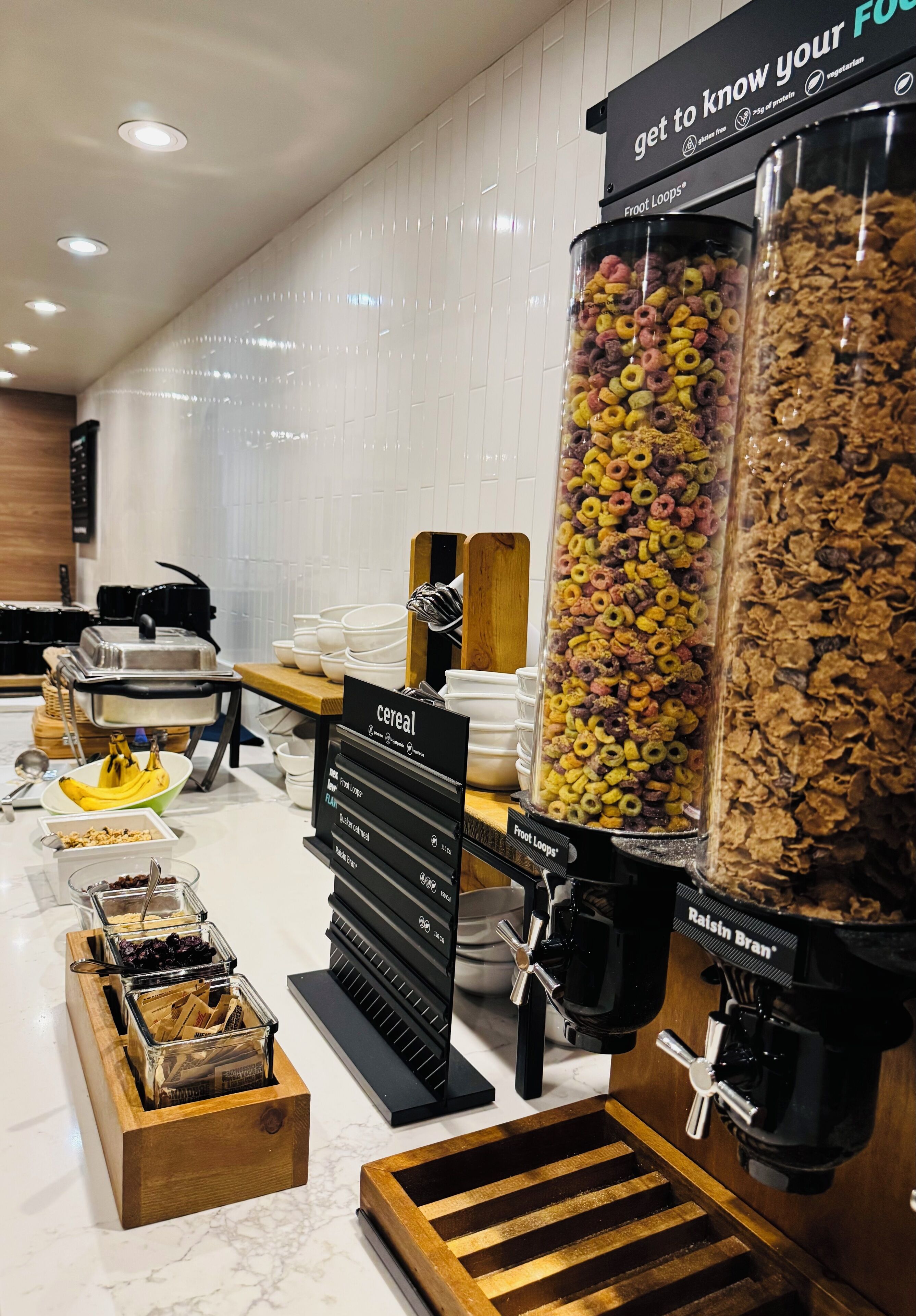 Un bufé de desayuno con diversos cereales, una máquina de café y un cartel que indica 'conoce tus alimentos'.