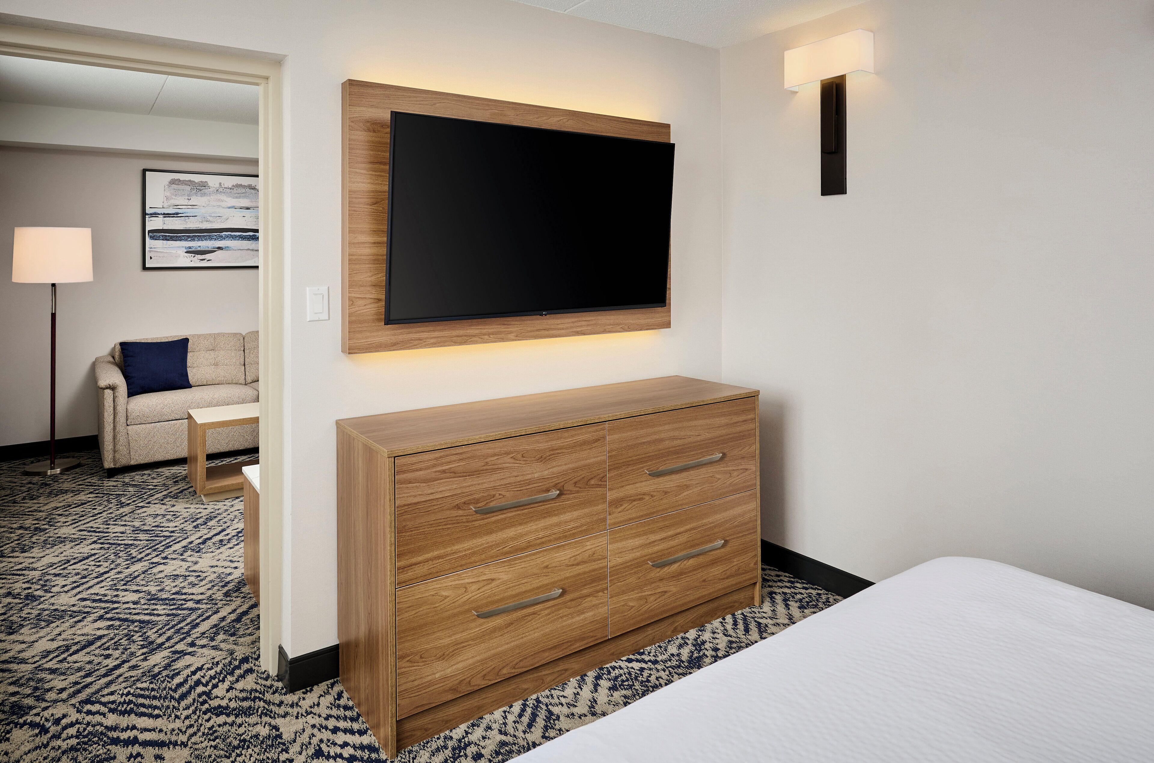 Hotellihuone, jossa on seinälle asennettu taulutelevisio, puinen lipasto laatikoineen, valkoisilla vuodevaatteilla varustettu sänky ja kuviollinen matto.