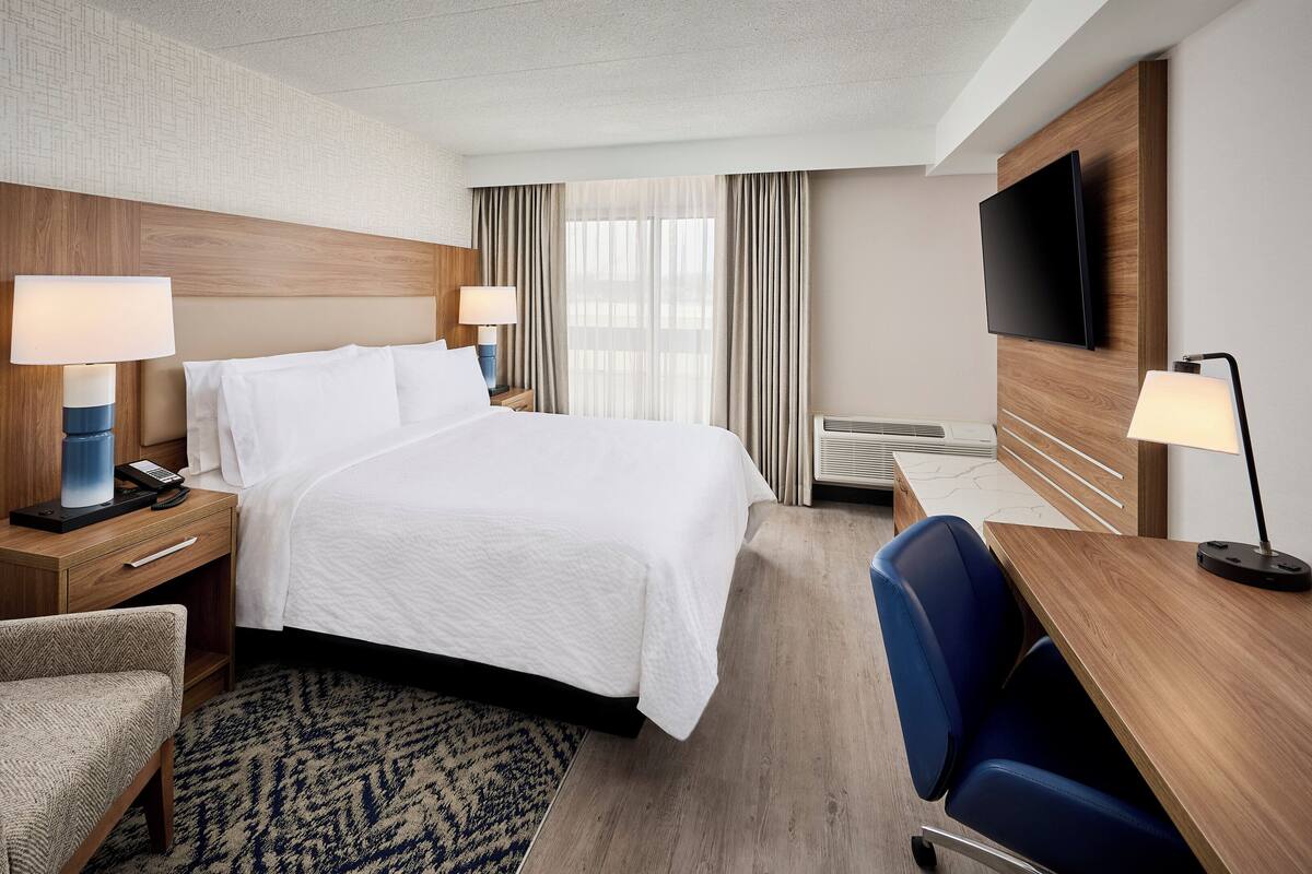 A hotel room with a bed, bedside tables with lamps, a desk with a chair, a TV, and a patterned rug.