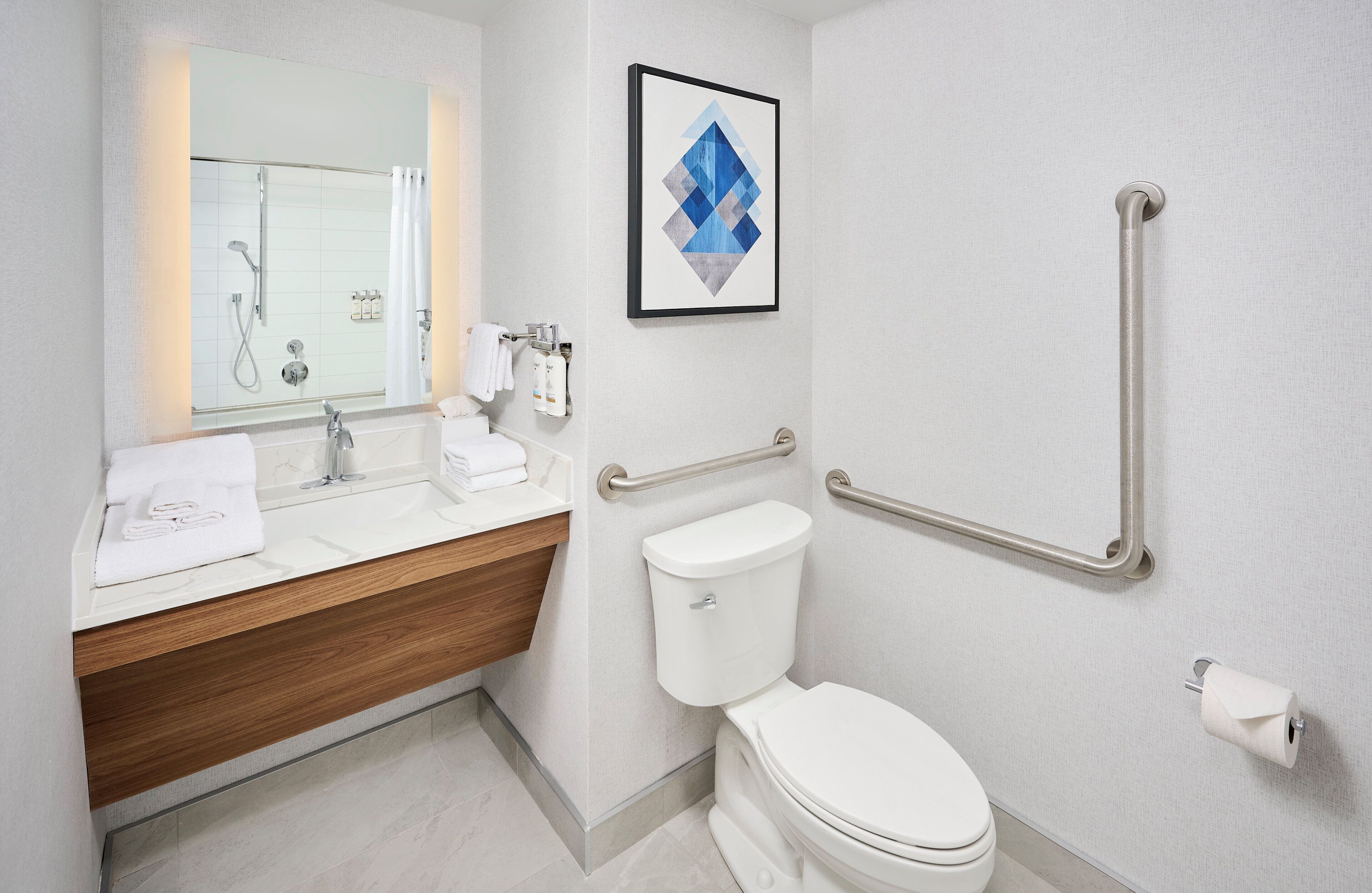 A modern bathroom with a white sink, mirror, and toilet. There is a framed artwork on the wall and a handrail on the right side.