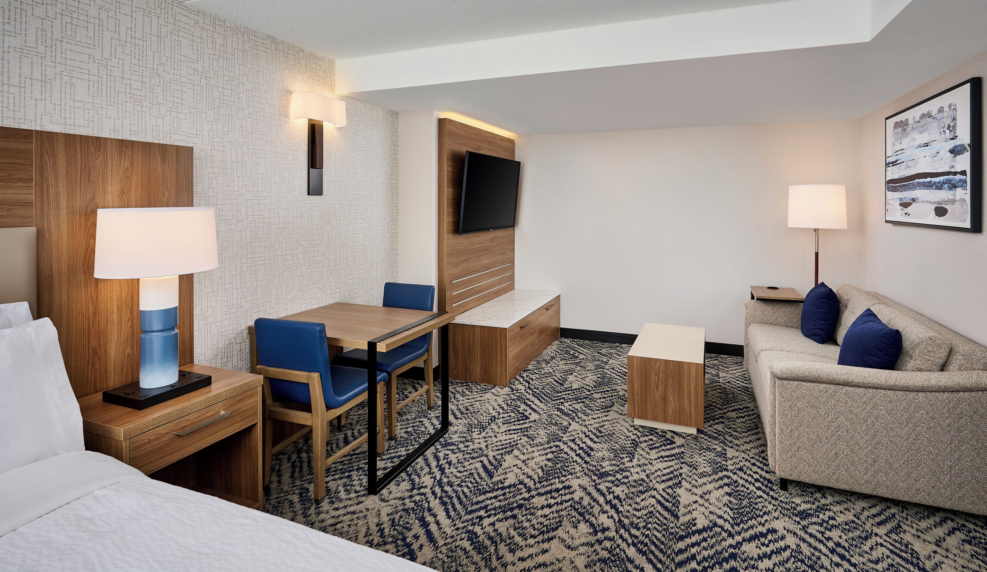 Habitación de hotel con cama, escritorio con silla, sofá, televisión y alfombra estampada.