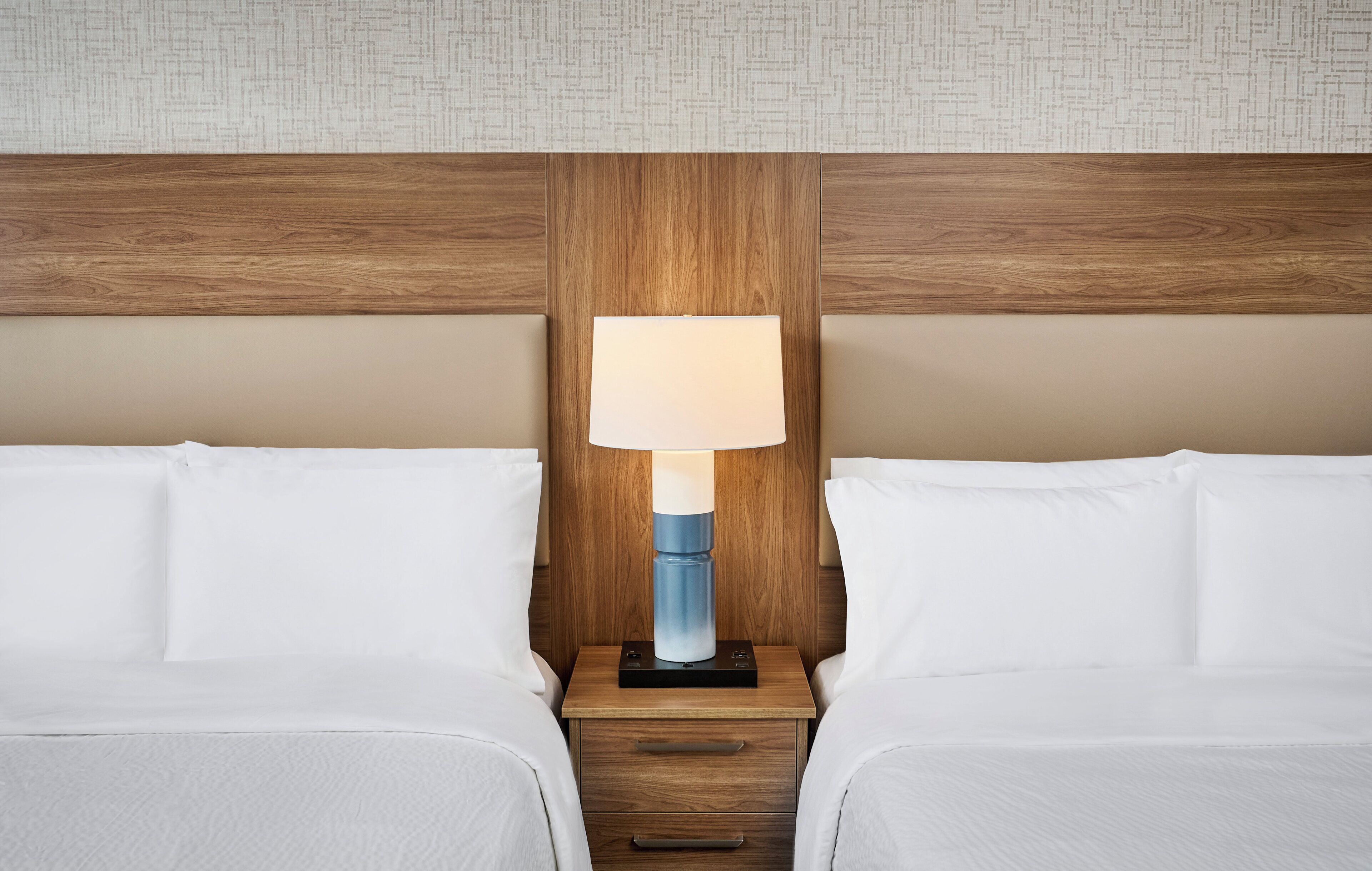 A hotel room with two beds, a wooden headboard, a nightstand with a lamp, and a plain wall with a subtle pattern.