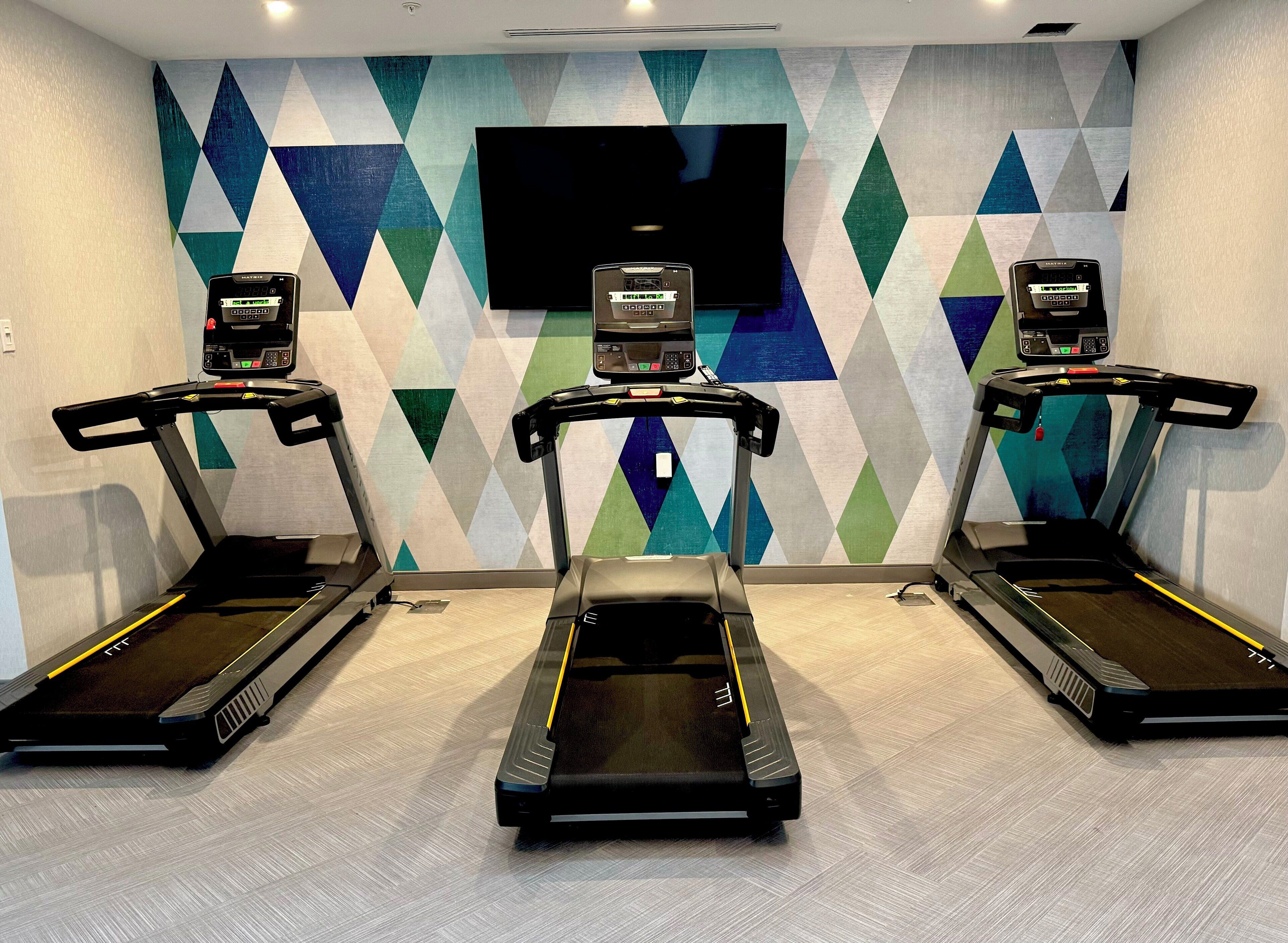 Une salle de sport équipée de trois tapis de course, d’un grand téléviseur fixé au mur et d’un mur décoré de motifs.