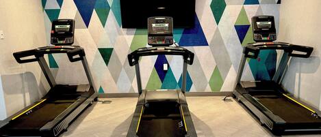 Une salle de sport équipée de trois tapis de course, d’un grand téléviseur fixé au mur et d’un mur décoré de motifs.