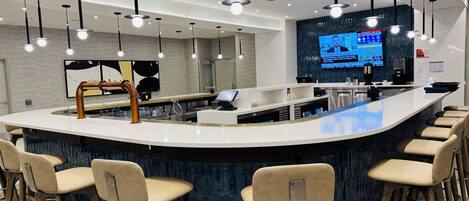 A modern bar area with a curved counter, bar stools, pendant lighting, a large flat-screen TV, and a coffee machine.