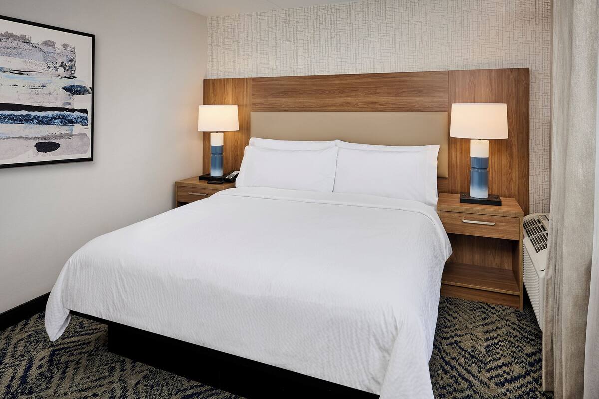 A neatly made bed with white linens, two bedside tables with lamps, a wall-mounted artwork, and a patterned carpet.