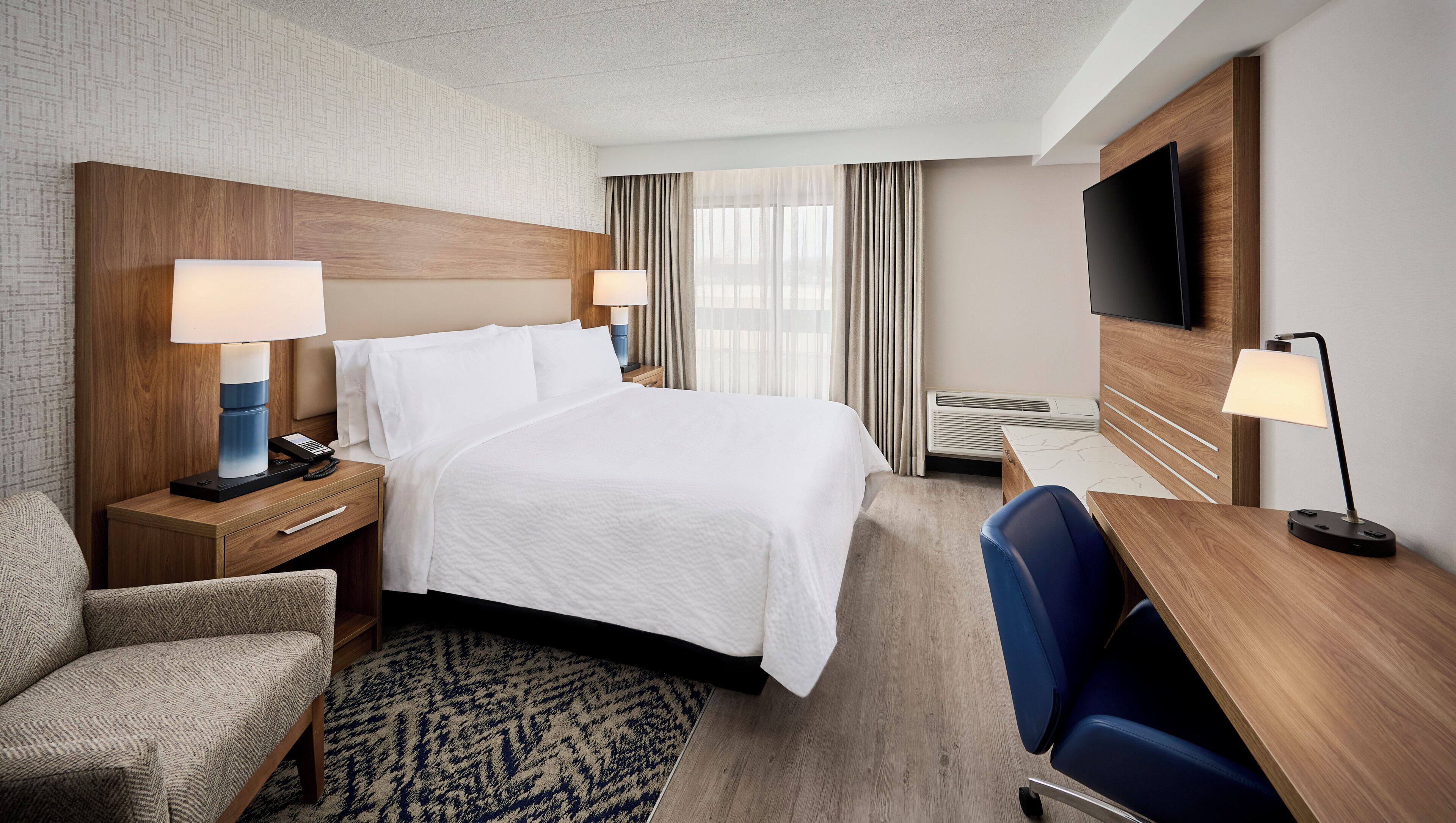 A hotel room with a bed, bedside tables with lamps, a desk with a chair, a TV mounted on the wall, and a patterned rug.