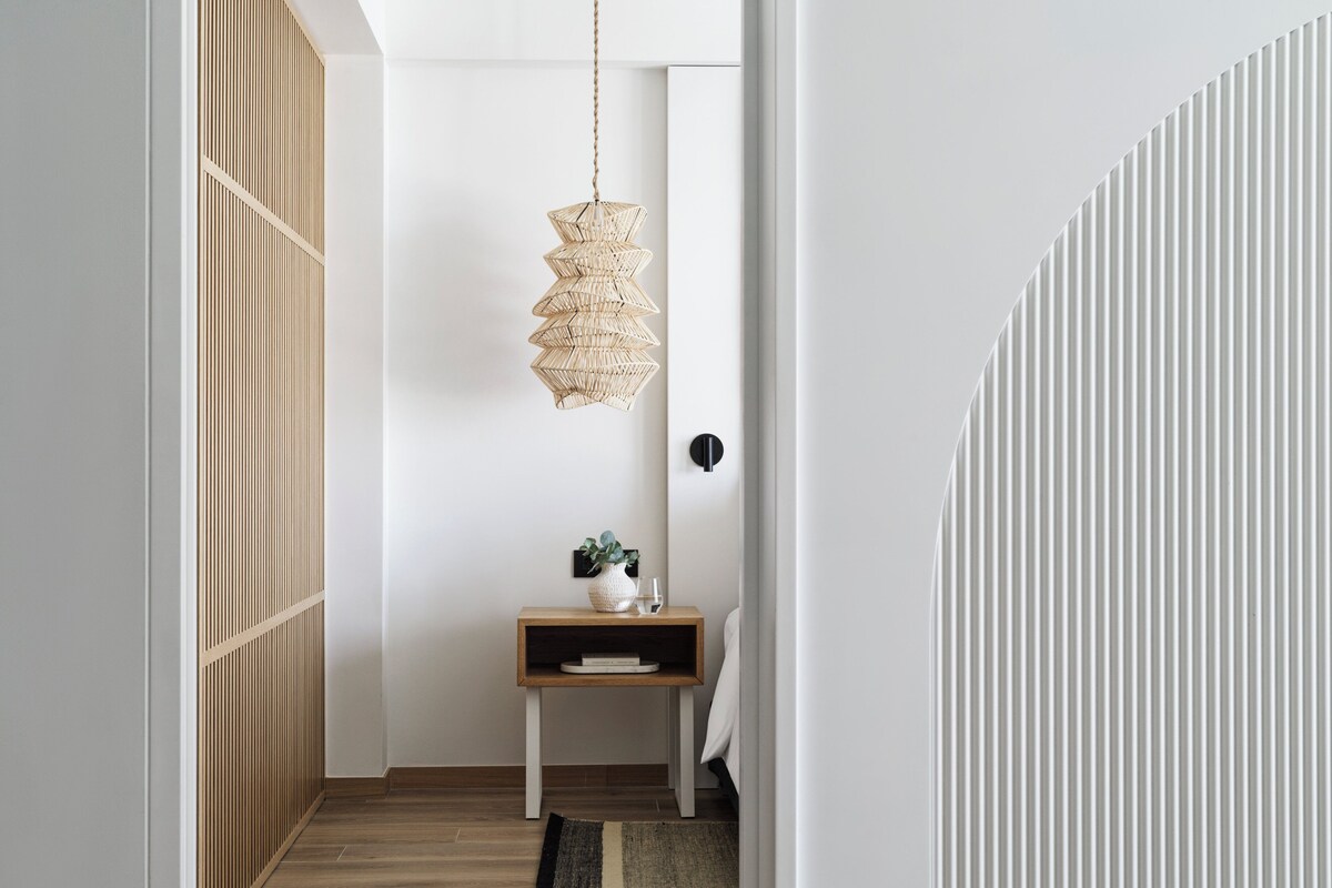 Un pasillo con suelo de madera, una lámpara de fibra natural, una pequeña mesita de madera con una planta y un radiador blanco.