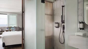 A modern hotel room with a large bed, a shower area with a handheld showerhead, and a bathroom with a marble wall and a large mirror.