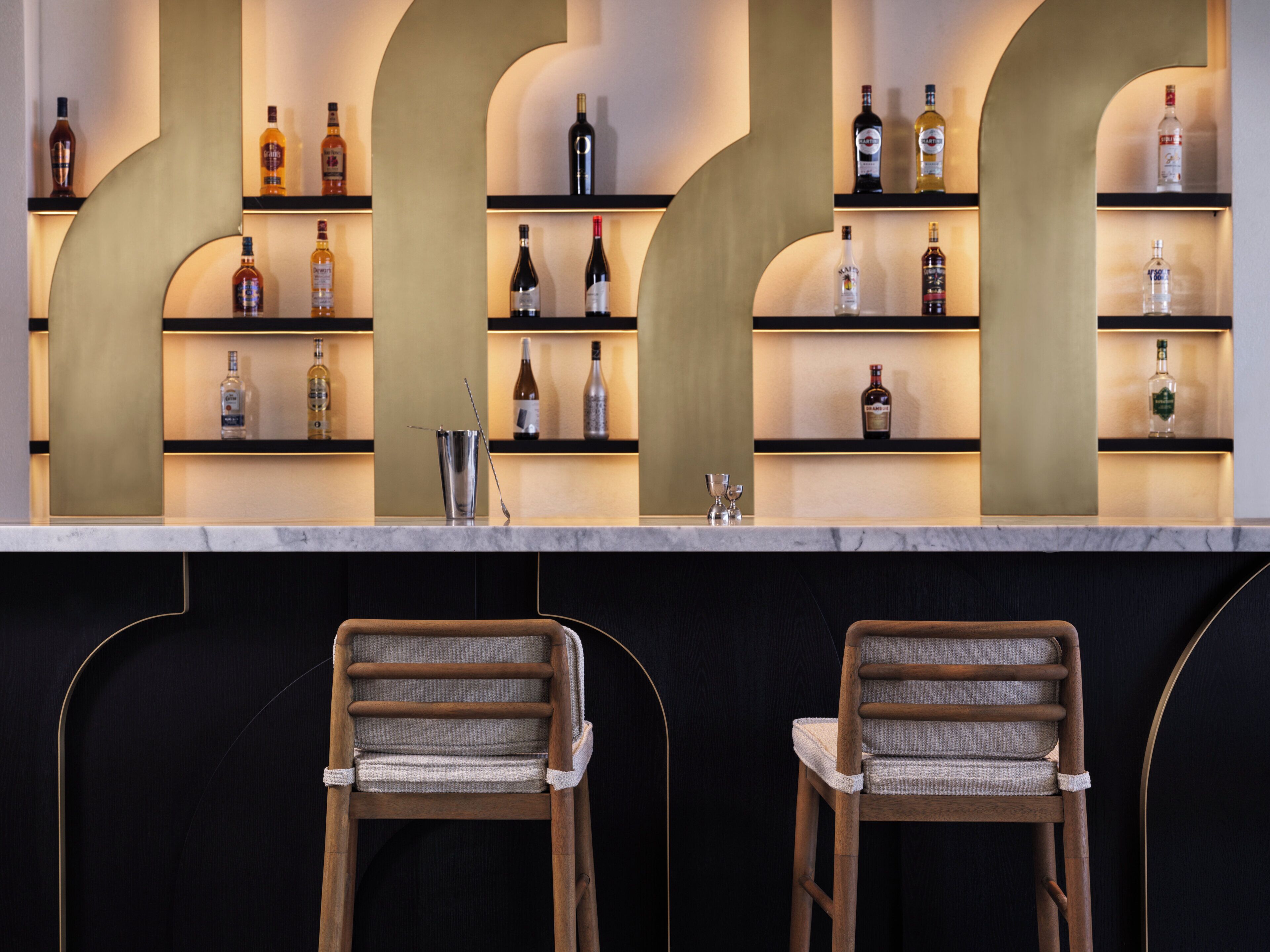 Un coin bar avec deux chaises en bois, un comptoir en marbre et des étagères remplies de différentes bouteilles d’alcool.