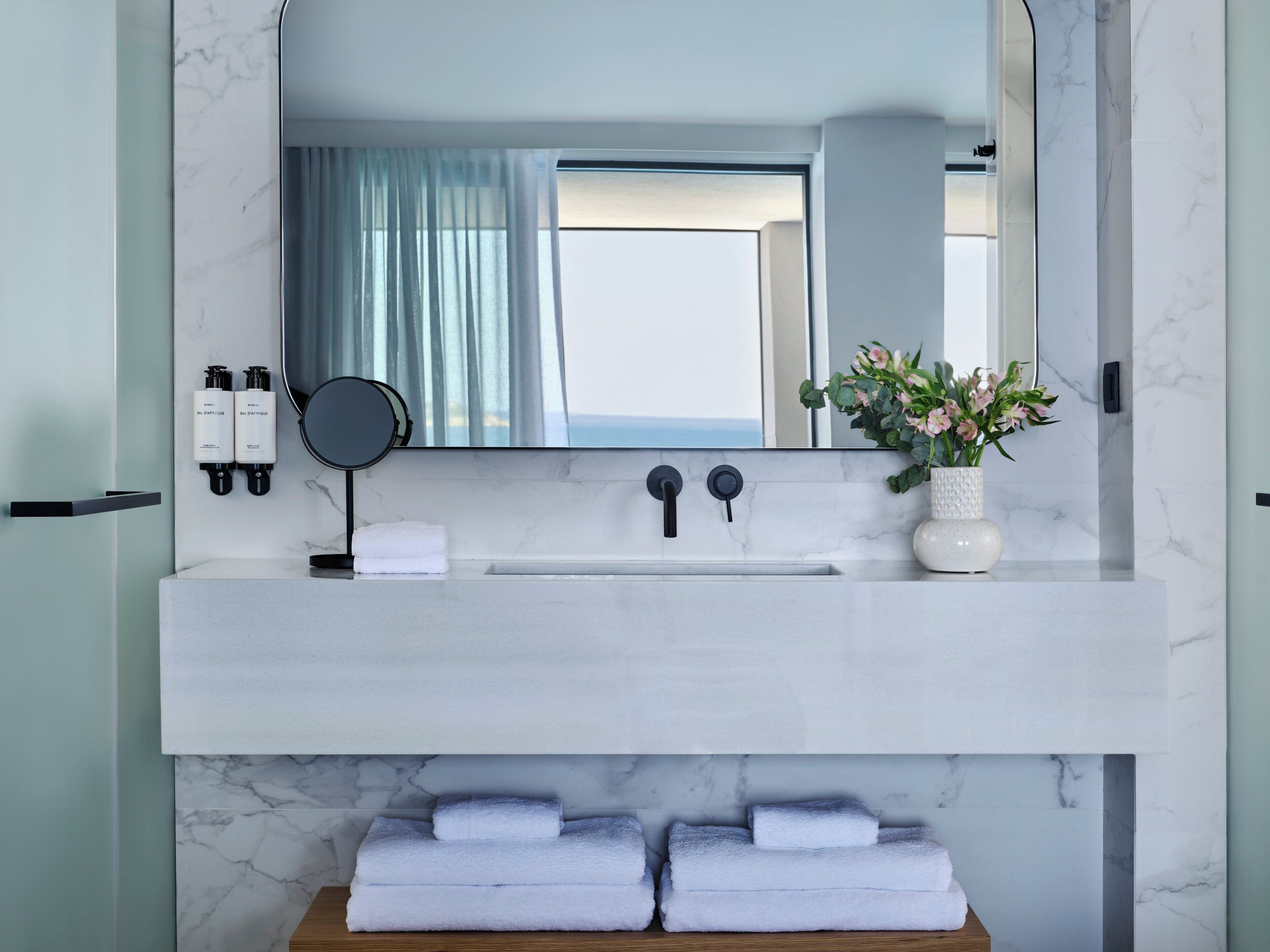A bathroom with a white marble sink, two black faucets, a round mirror, a towel rack with folded towels, and a vase with flowers.