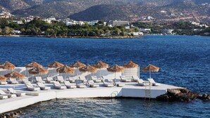 Une station balnéaire dotée de chaises longues et de parasols en chaume, d’une piscine, et offrant une vue sur la mer et les montagnes.