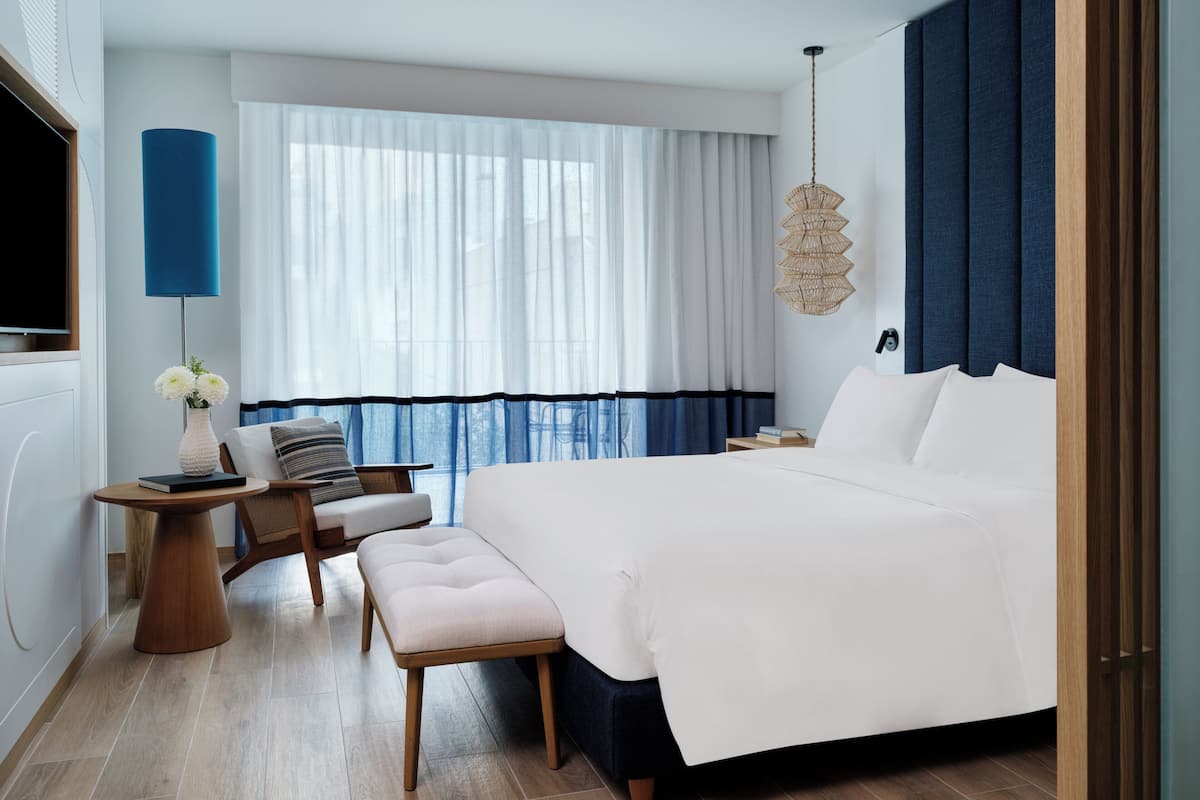 Ein modernes Hotelzimmer mit einem großen Bett, einem Schreibtisch mit Stuhl, einem kleinen Tisch und einem großen Fenster mit Vorhängen.