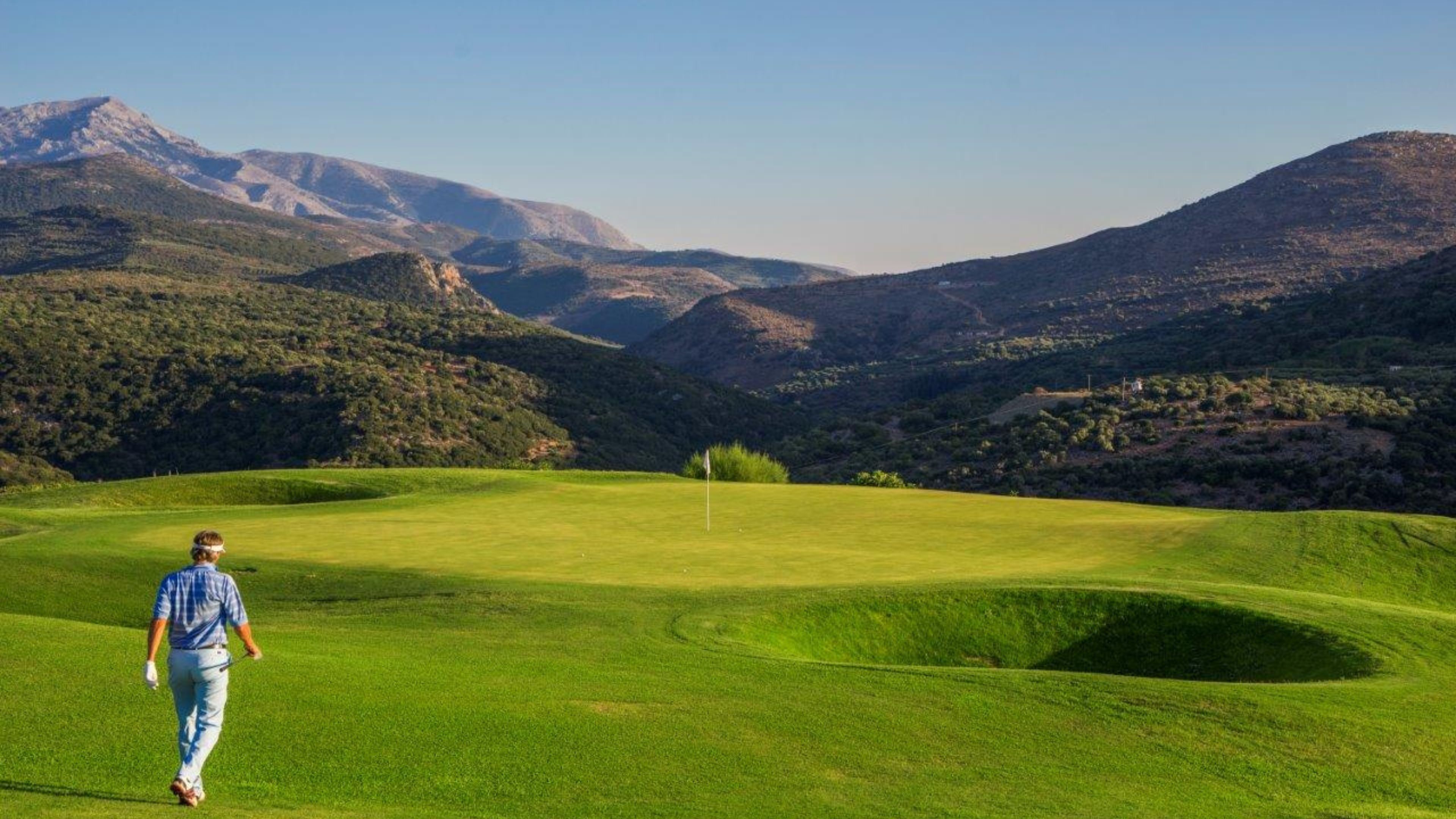 Un campo de golf con una persona caminando sobre el césped, rodeado de montañas.