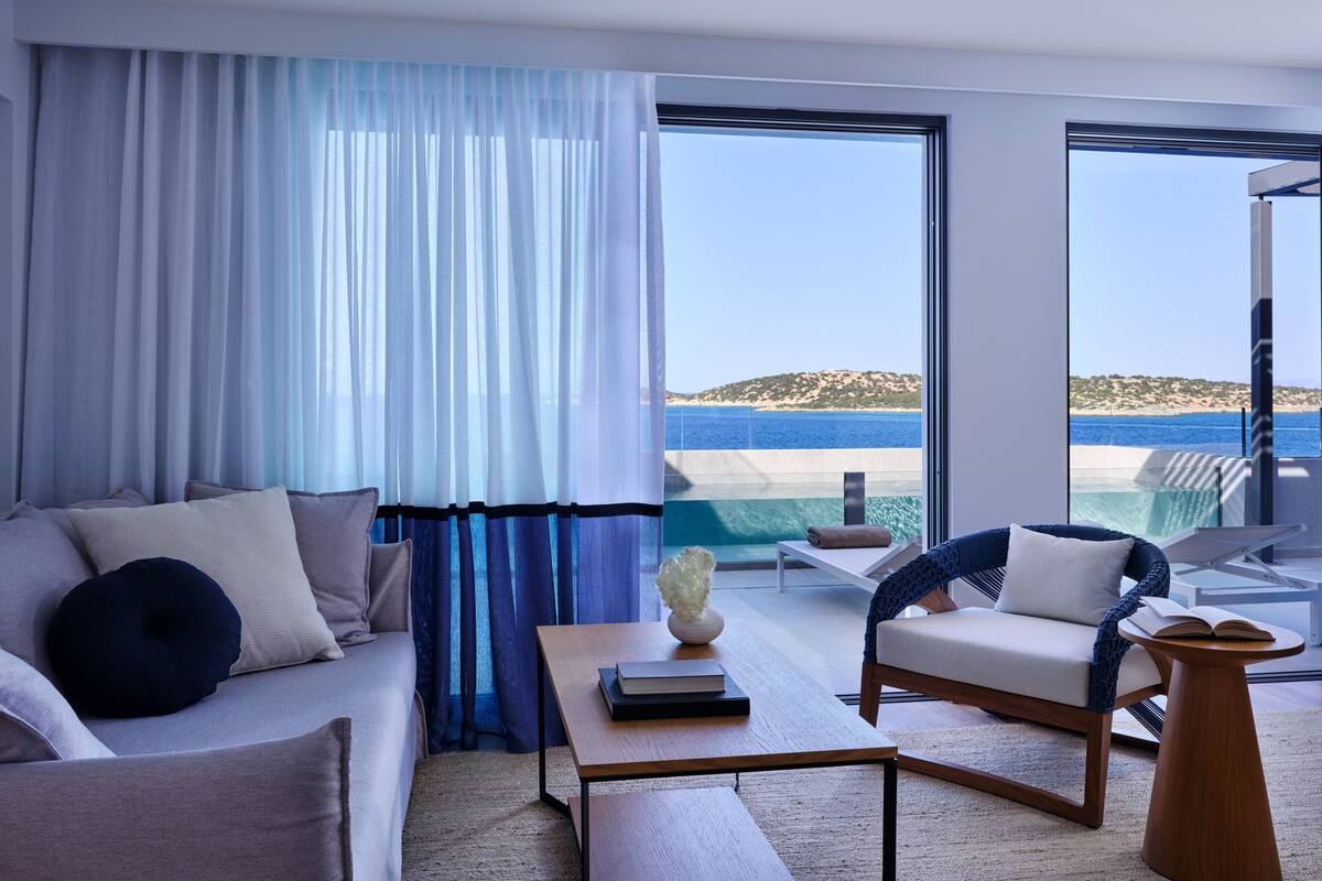 Ein modernes Hotelzimmer mit großem Fenster und Meerblick, inklusive Strand. Es gibt ein Sofa, einen Sessel und einen kleinen Tisch mit einem Buch und einer Vase.