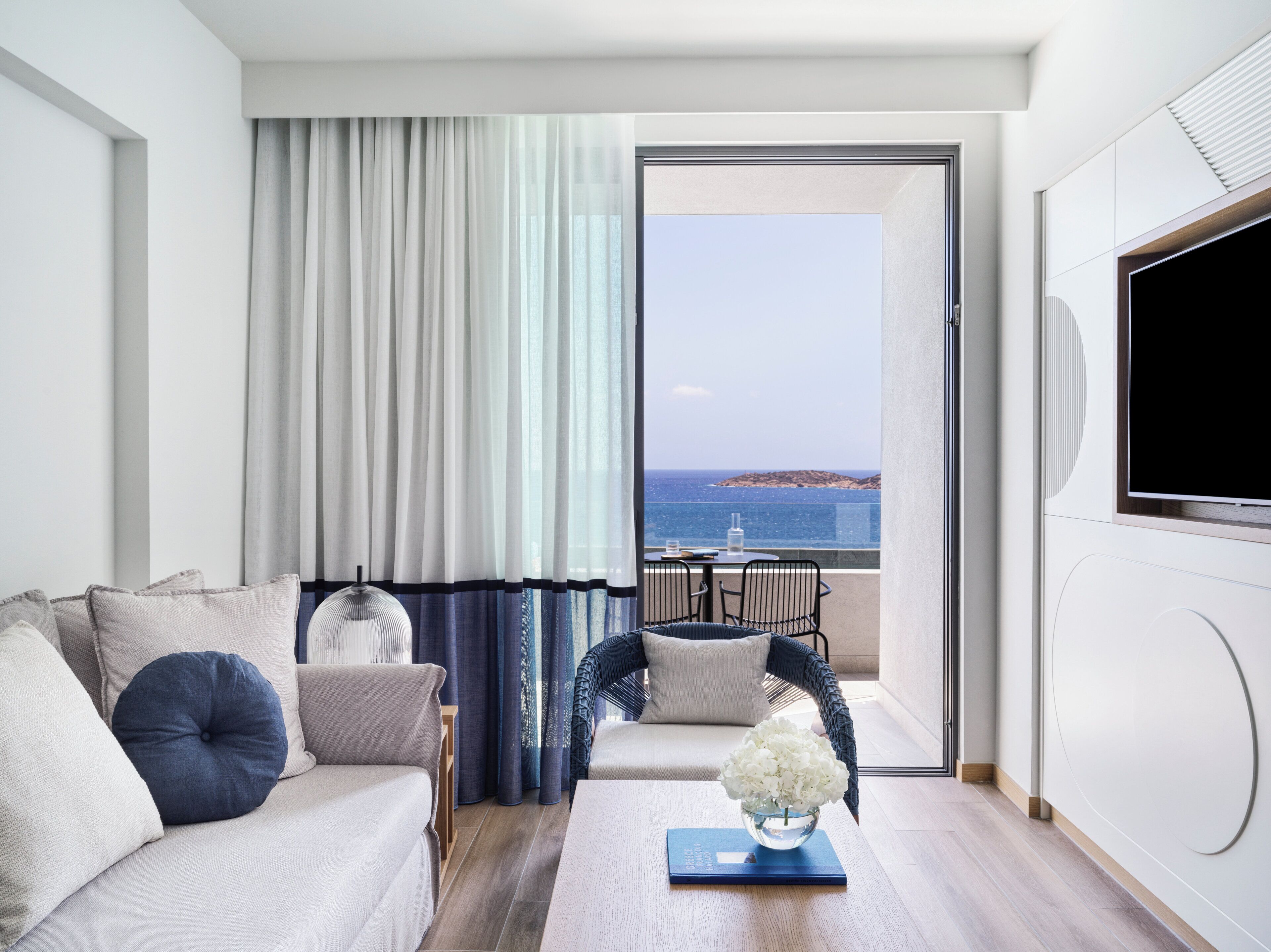 Una habitación de hotel moderna con sofá, una mesa de centro con un florero, vista al mar y un televisor grande en la pared.