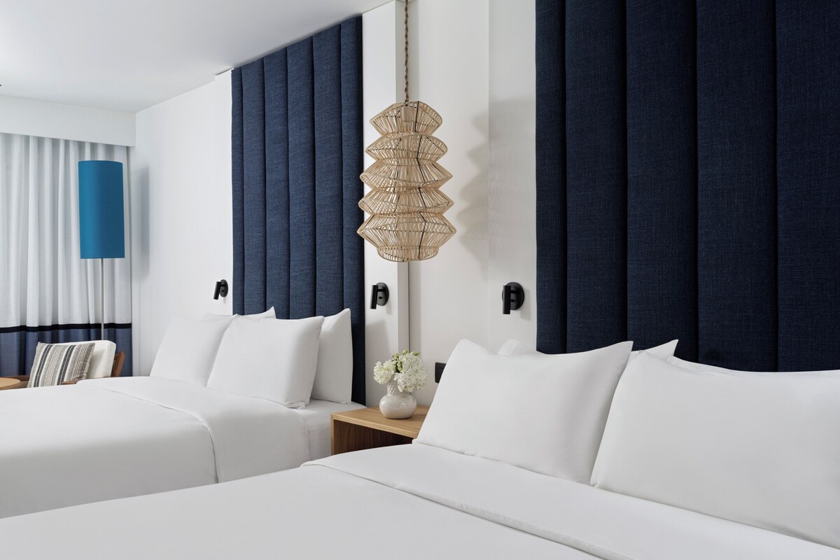 Habitación de hotel con dos camas, un ventanal con cortinas, una lámpara de pared y una mesita con un florero.