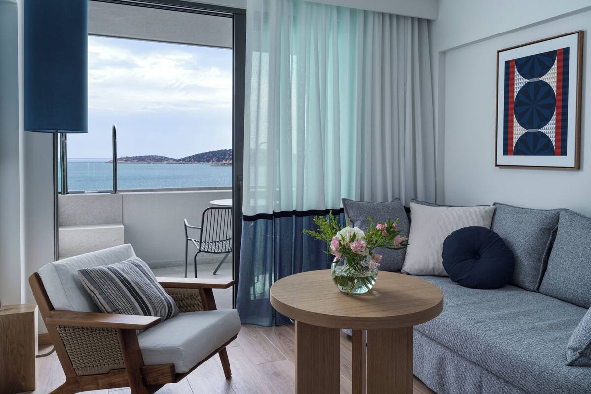 A modern living room with a sofa, a chair, a small table with a vase of flowers, a view of the sea through large windows, and a framed abstract painting on the wall.