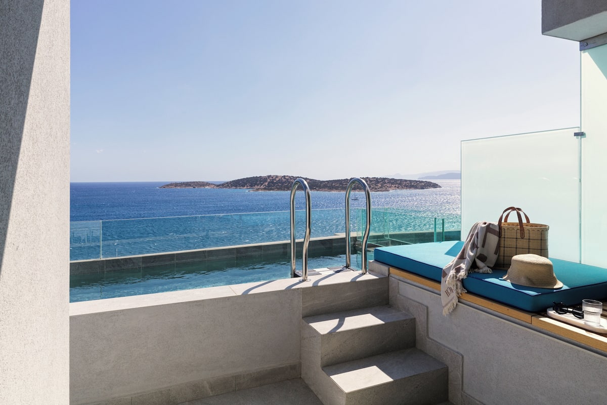 Ein Balkon mit Pool und Meerblick, eine Treppe sowie eine Sitzecke mit Hut und Sonnenbrille.
