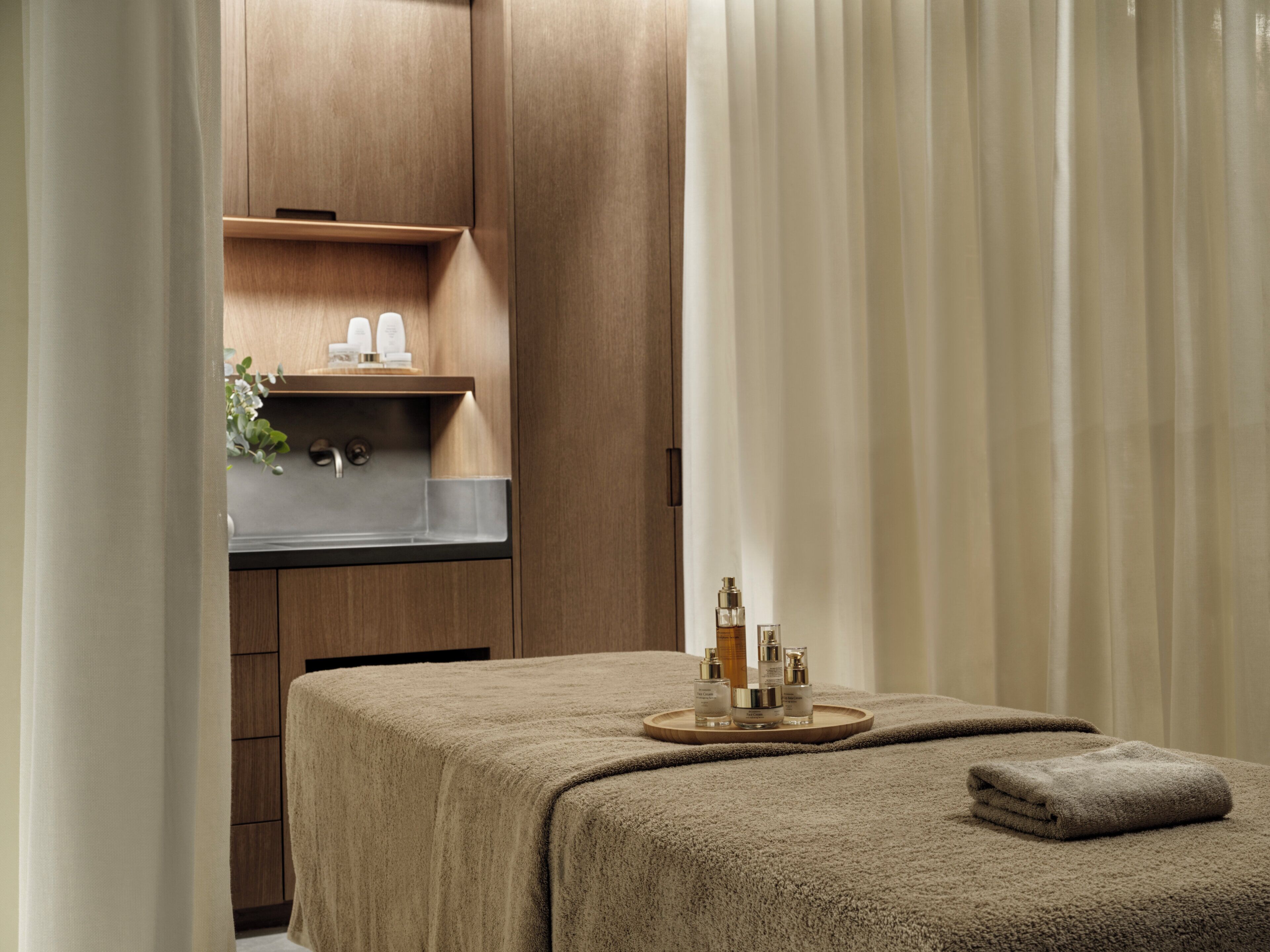 A spa room with a massage table, a wooden shelf with toiletries, and a large window with curtains.