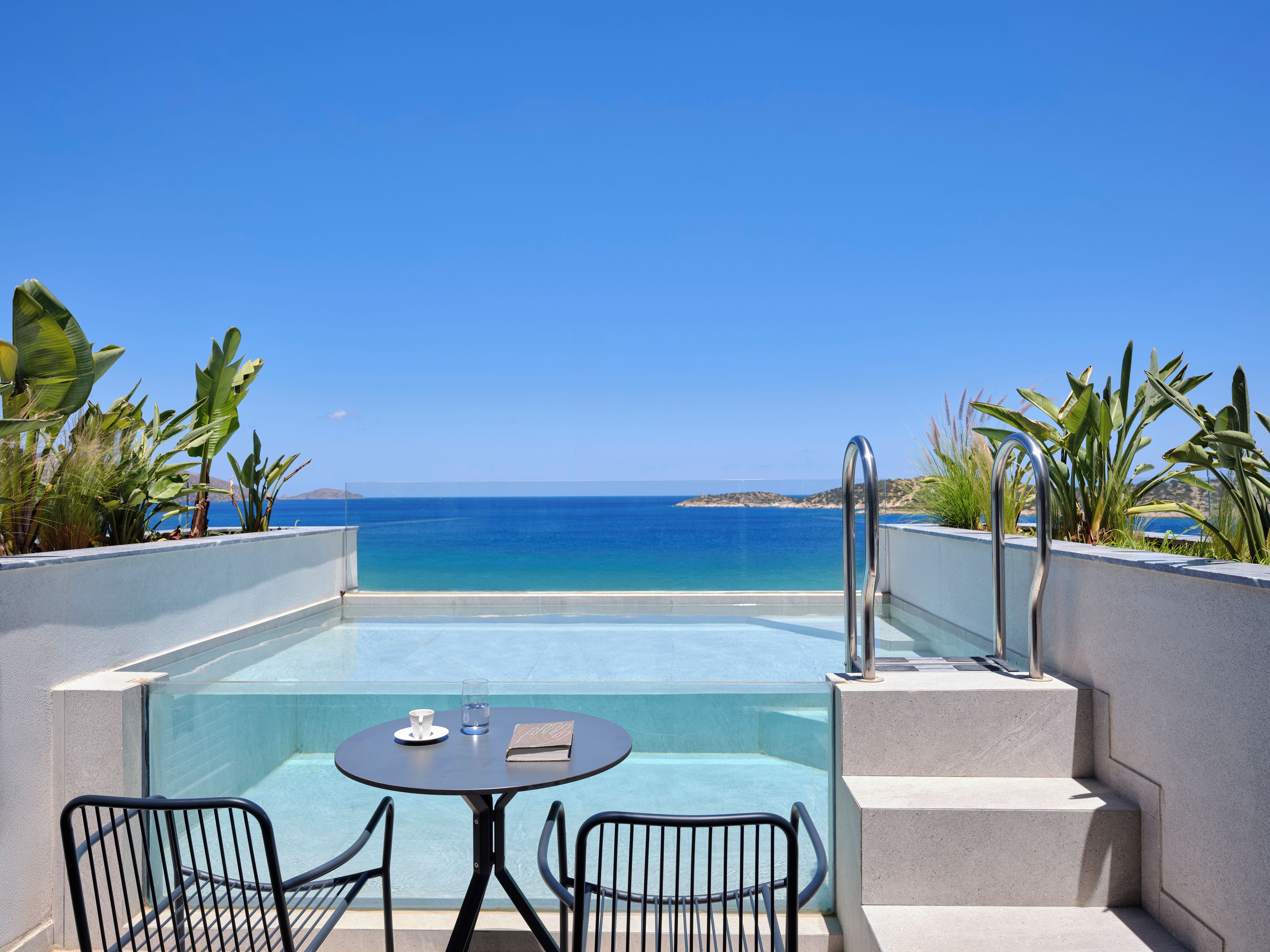 A pool with a view of the sea, a table with chairs, and a book.