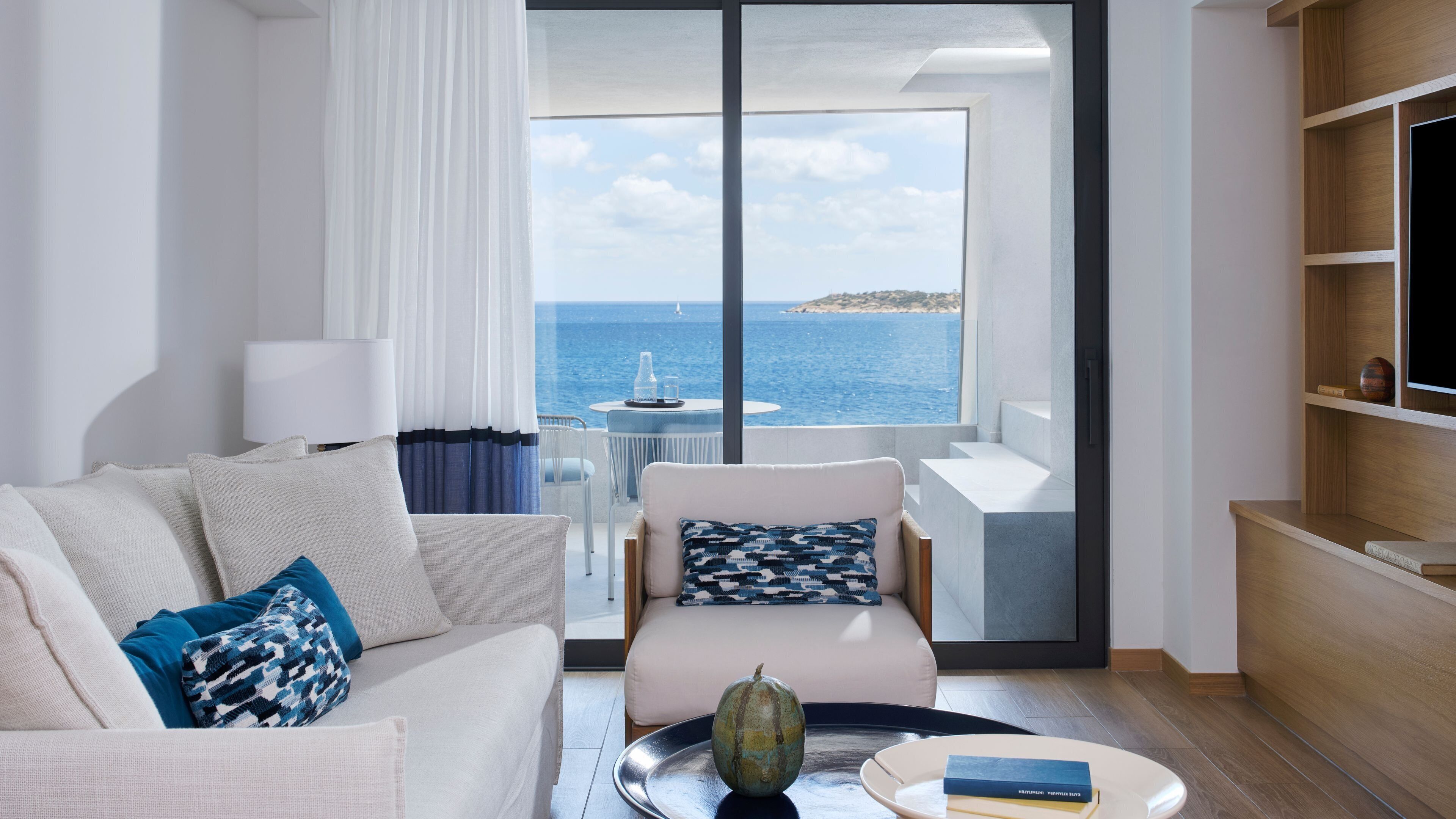 A modern living room with a view of the sea, a sofa, a chair, a coffee table, and a bookshelf.