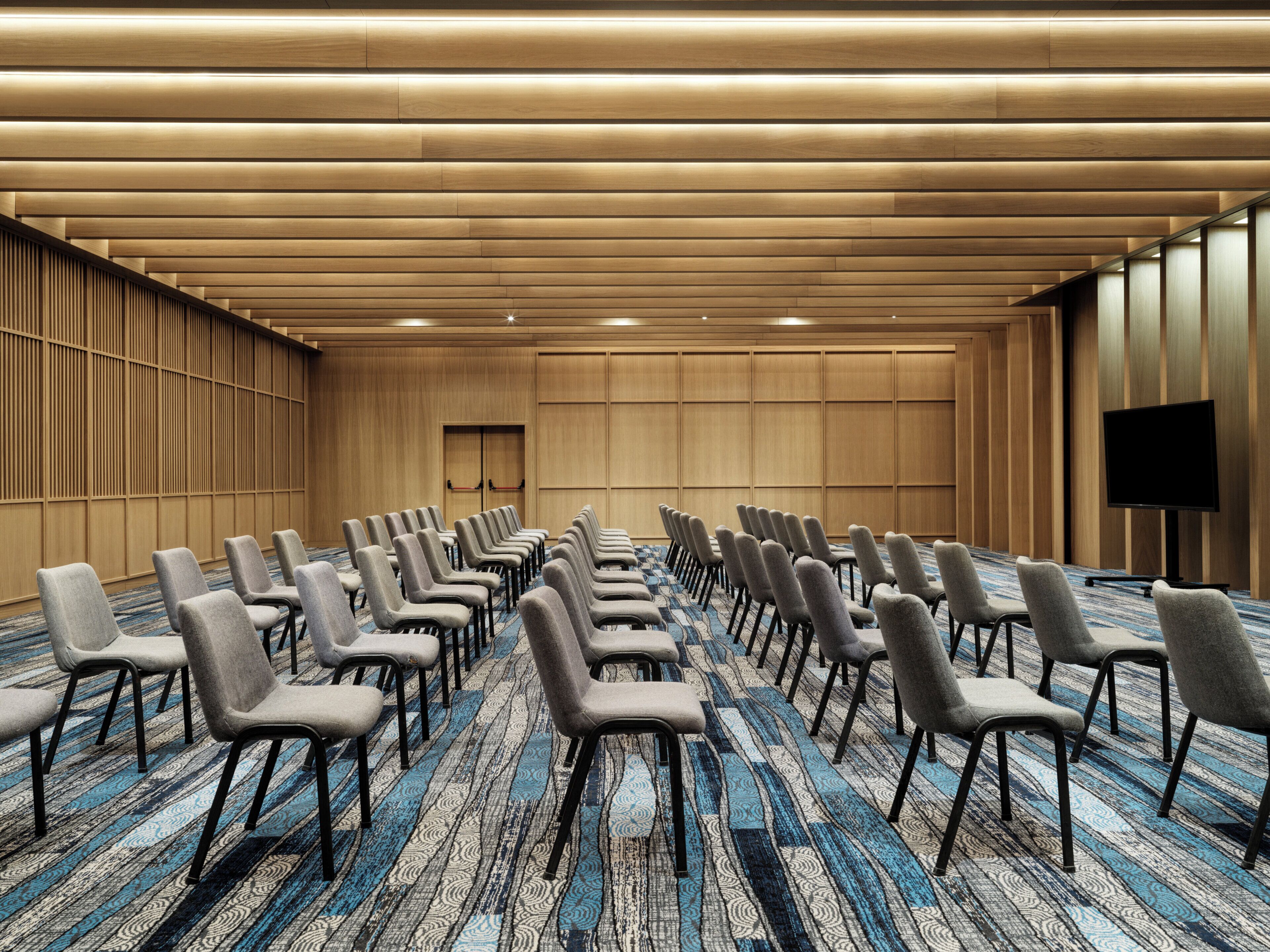 A conference room with rows of chairs, a carpeted floor, wooden panel walls, and a television on the right.