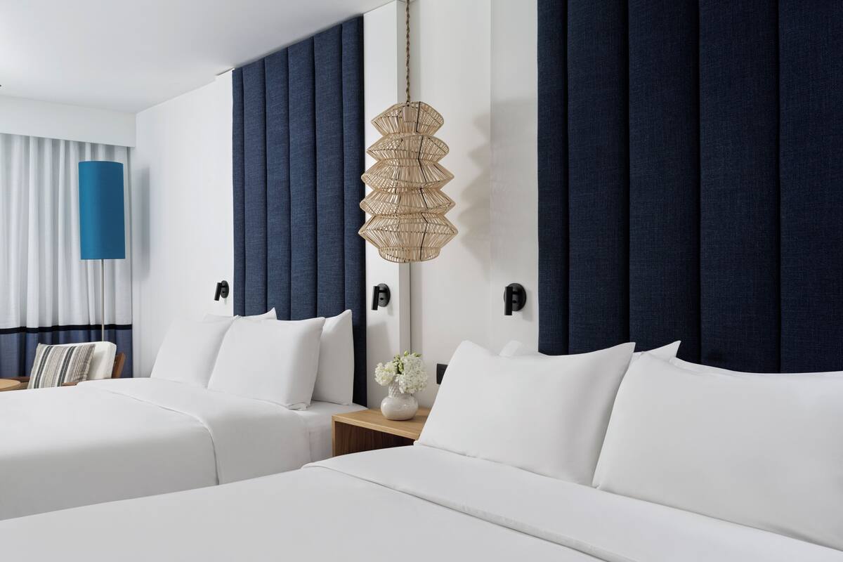 Ein Hotelzimmer mit zwei Betten, einem großen Fenster mit Vorhängen, einer Wandlampe und einem kleinen Tisch mit einer Vase.