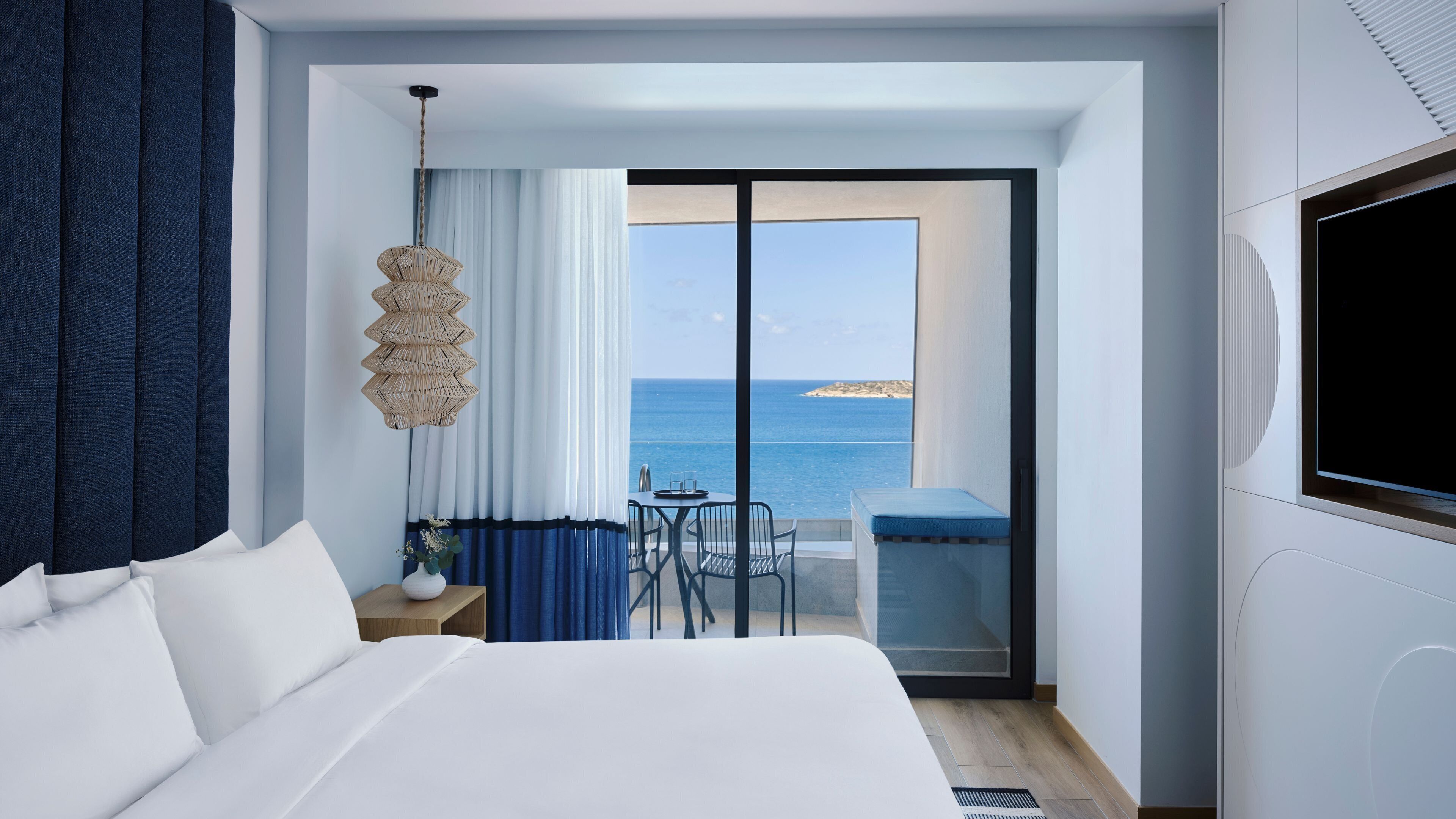 Habitación de hotel con cama, una pequeña mesa con sillas, lámpara colgante y vistas al mar.