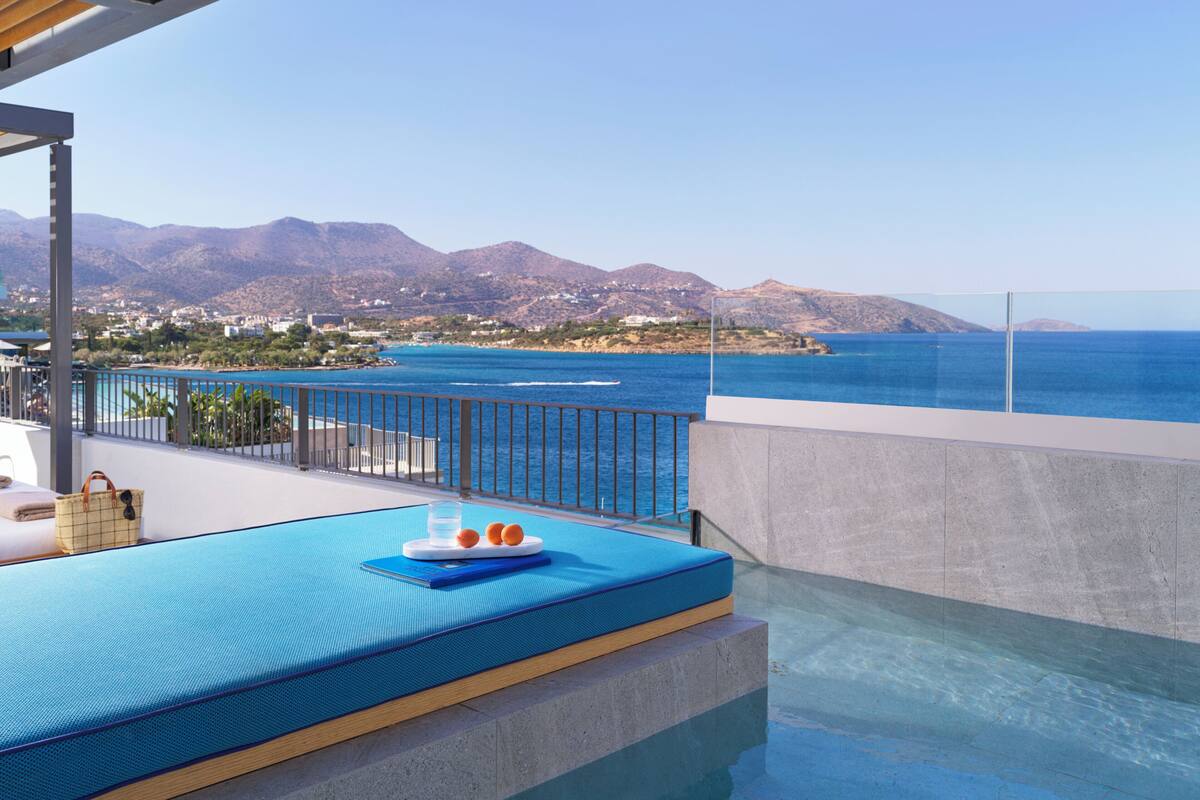 Ein Poolbereich mit Blick auf das Meer und die Berge, eine Liege und ein Tisch mit Getränken und Snacks.