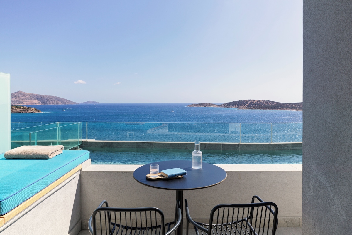 Un balcón con vista al mar, una mesa con un libro y un vaso, y una toalla en el asiento.