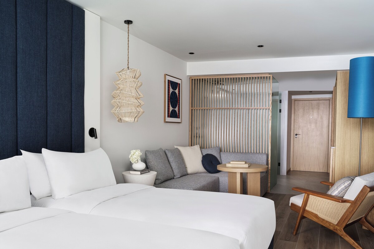 Habitación de hotel moderna con una cama grande, un sofá, una mesita redonda pequeña, una silla y un ventanal amplio.
