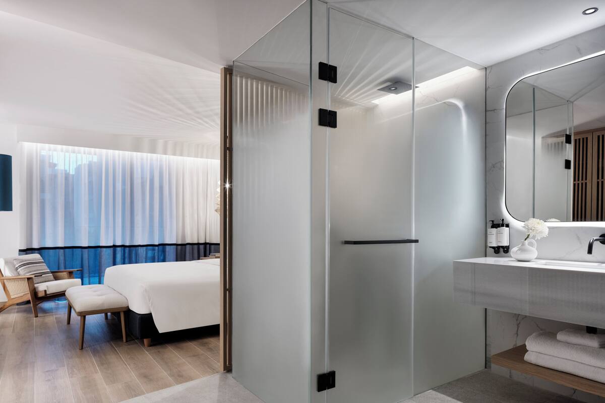 Ein modernes Hotelzimmer mit Glastrennwand, einem Bett, einer Sitzecke mit einem Sessel, einem großen Spiegel und einem Badezimmer mit Waschbecken und Handtuchhalter.