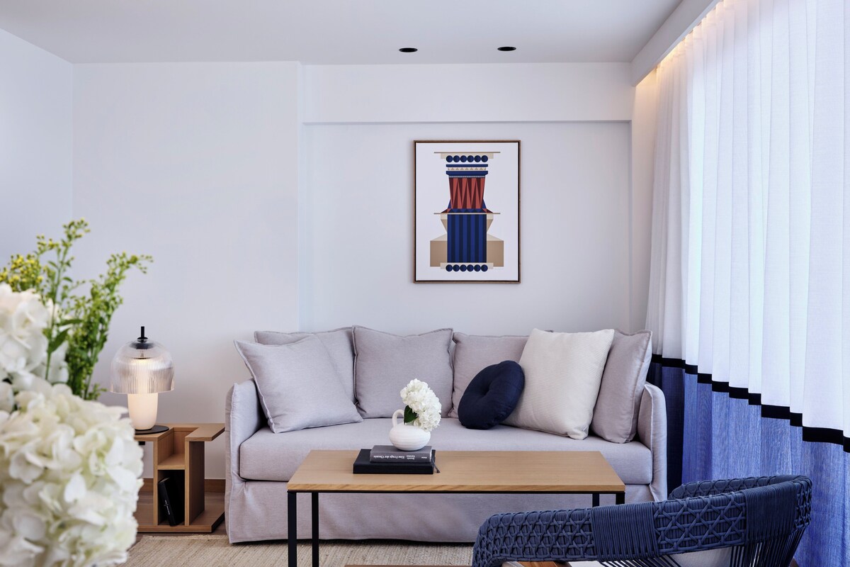 Un moderno salón con un sofá, una mesa de centro y una silla. Hay una obra de arte enmarcada en la pared y una lámpara sobre una mesita auxiliar.