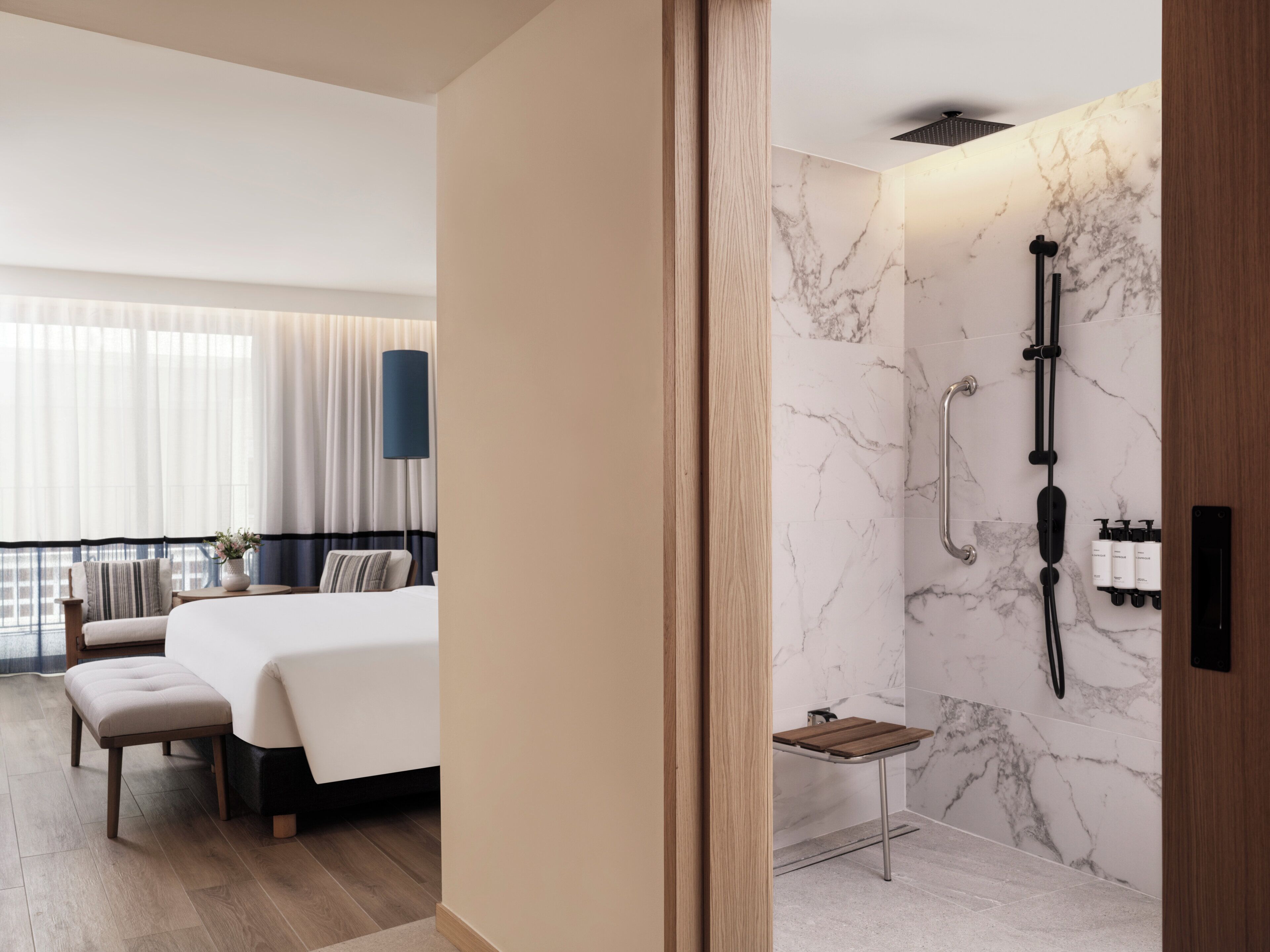 A modern hotel room with a large bed, a bedside table, a small bench, a bathroom with a shower, and a view of the city through large windows.