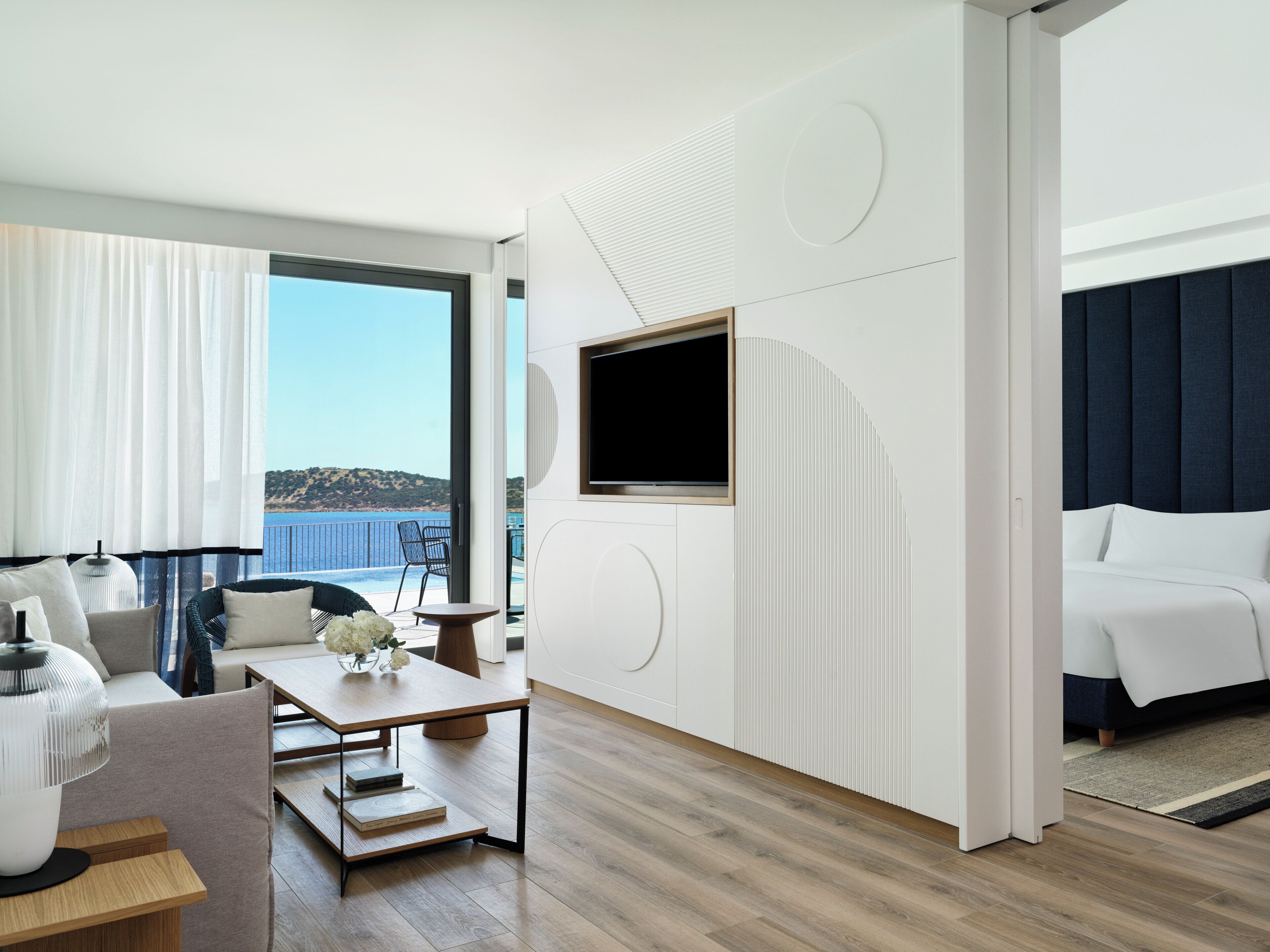 A modern hotel room with a balcony, a large TV, a bed, and a seating area with a table and chairs.