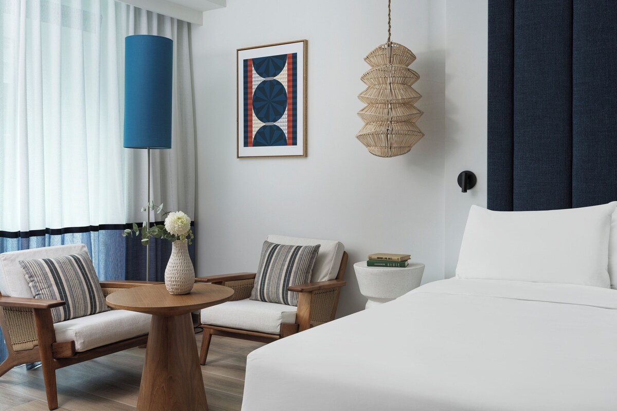 Habitación de hotel con una cama, dos sillones, una mesa redonda de madera, un florero con flores, un cuadro abstracto enmarco, una lámpara colgante de fibras tejidas y una mesita de noche con libros.