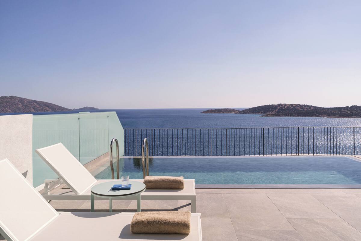 Ein Poolbereich mit Liegestühlen und einem Tisch, mit Blick auf das klare, blaue Meer und ferne Hügel.