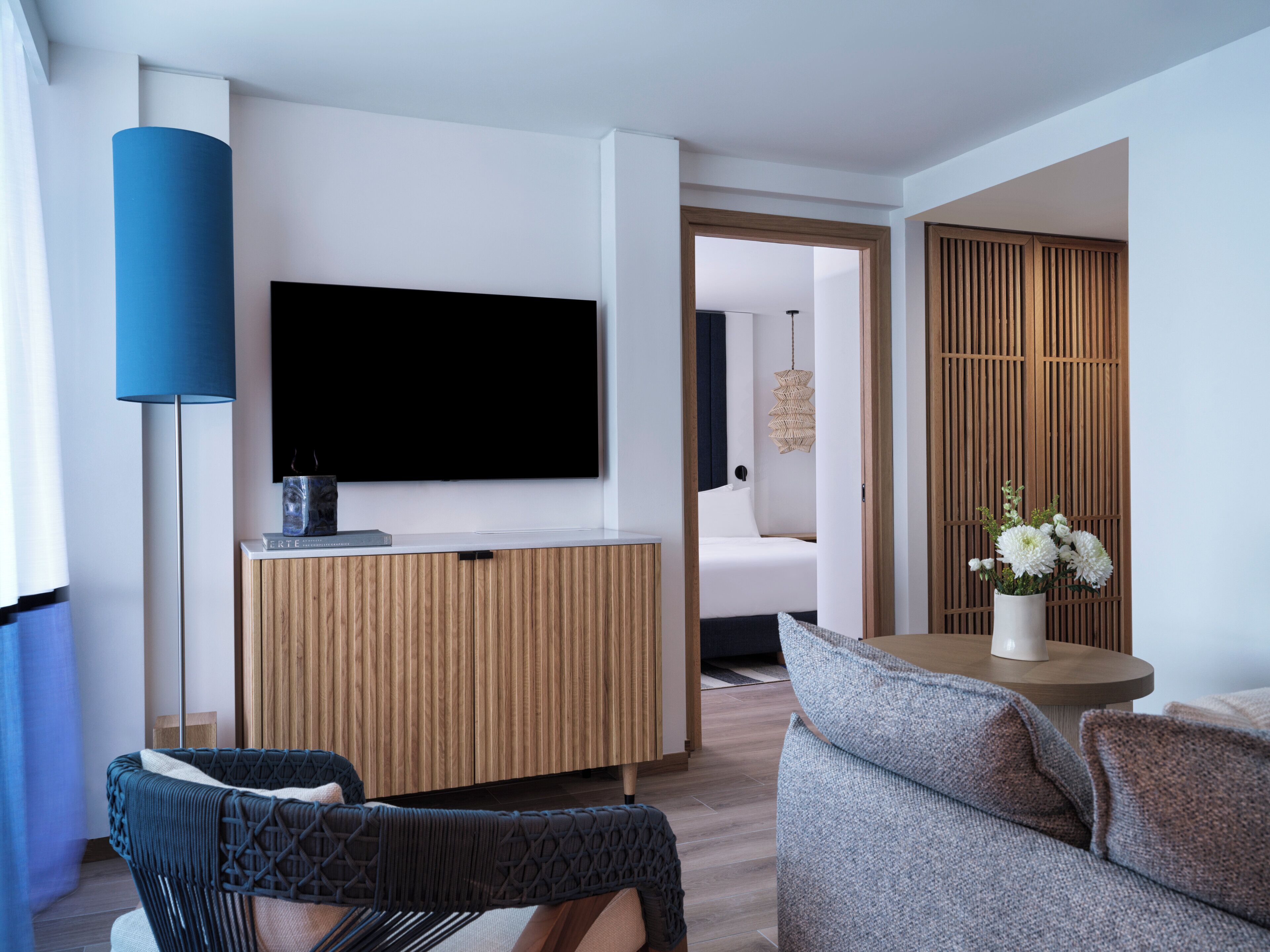 Ein modernes Hotelzimmer mit Flachbildfernseher, einer Holzkonsole, einer blauen Stehlampe, einem grauen Sofa und einem kleinen Tisch mit Blumenvase.