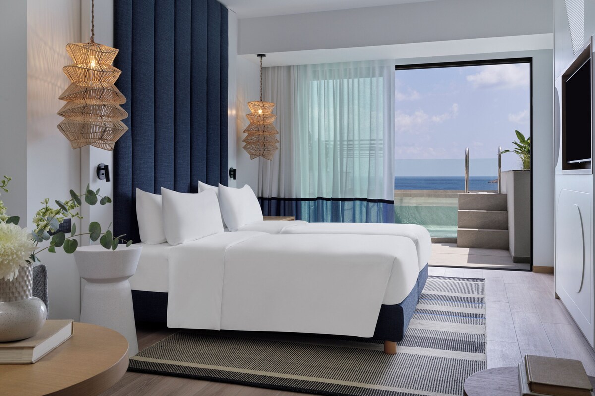 Habitación de hotel moderna con una cama grande, dos lámparas de noche, vista al océano y un balcón.