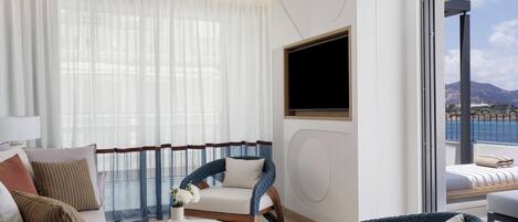 A modern hotel room with a sofa, armchairs, a small table, a TV, and a balcony with a view of the sea.