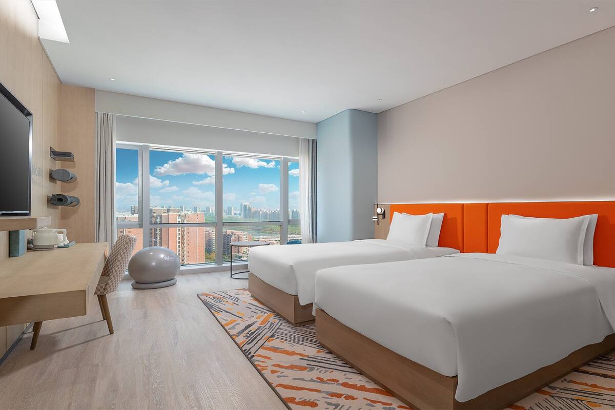 A modern hotel room with two beds, a desk, a TV, and a large window offering a city view.