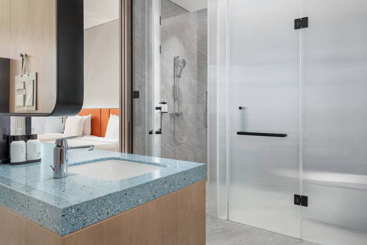 A modern bathroom with a granite countertop, a shower with a glass door, and a bed with orange headboard.