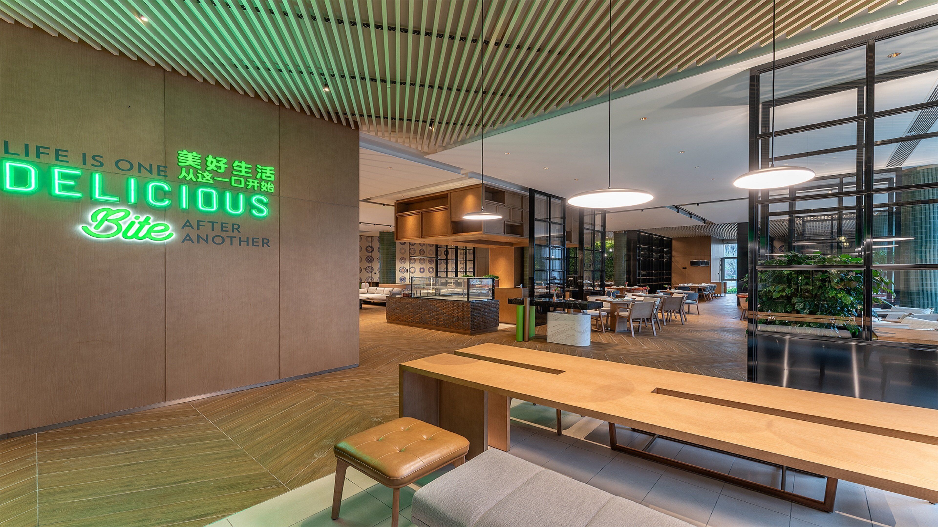 Un intérieur moderne avec un banc et des tables en bois, un panneau néon sur lequel on peut lire « UNE DÉLICE APRÈS L’AUTRE », et une vue sur l’extérieur grâce à de grandes fenêtres.