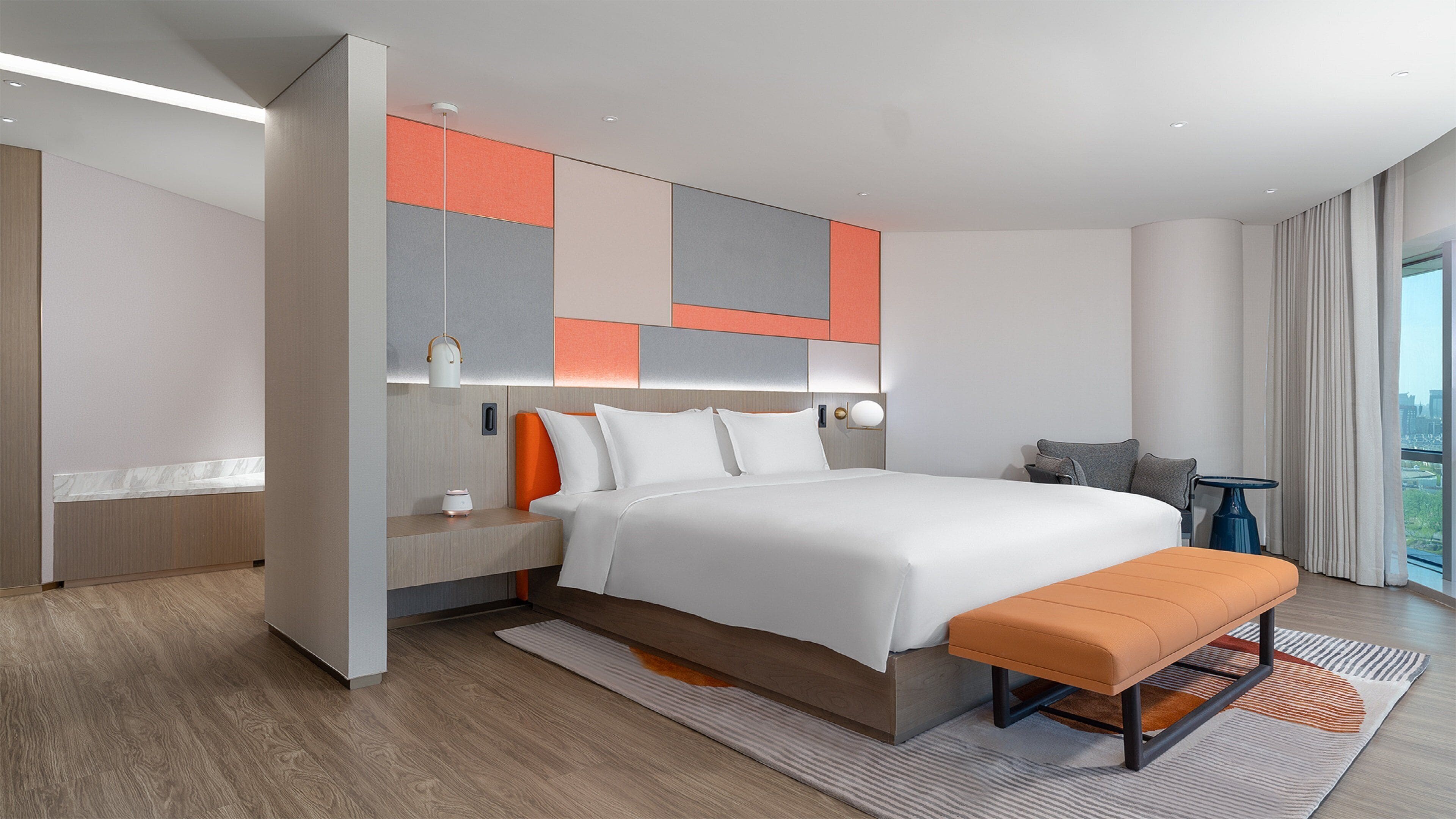 A modern hotel room with a large bed, a bench, a small table, and a view of the city.